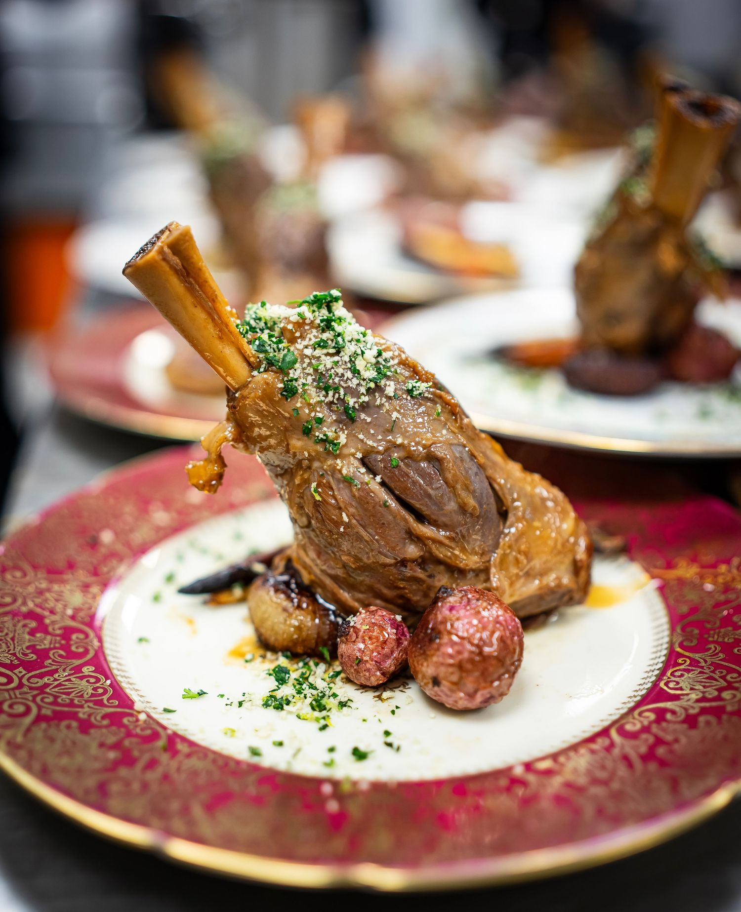 A succulent lamb shank with a bone, garnished with parsley and served with roasted red potatoes, presented on an ornate red  rimmed plate.