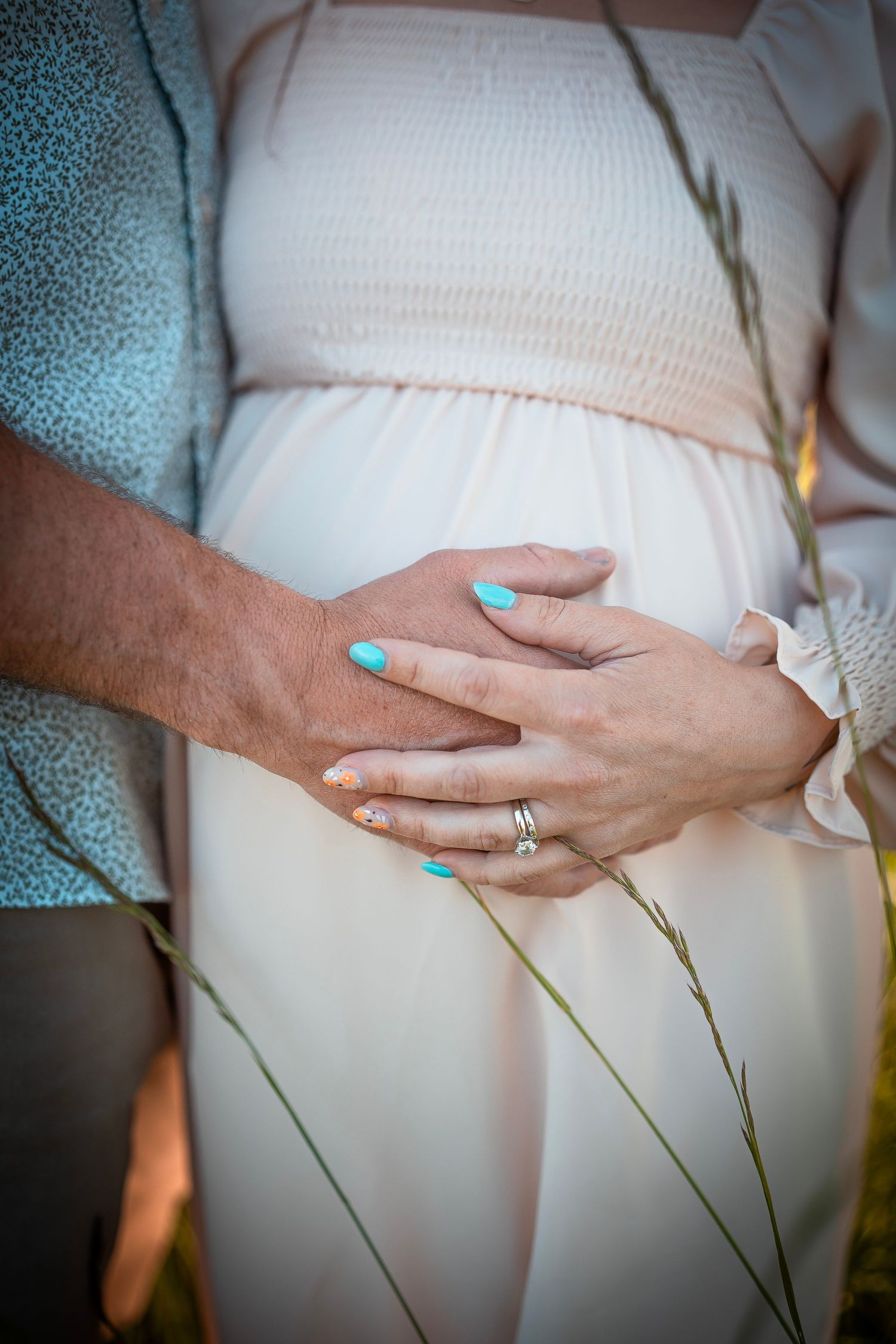 A pregnant woman holding her stomach. Man is also holding her stomach. She has light blue nails and light pink flowy dress.