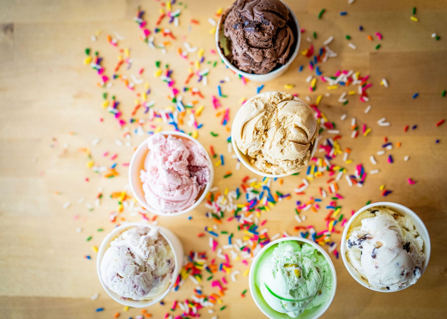 Photo of multiple ice-creams cups including chocolate, strawberry, mint chocolate chip, cookie dough, and vanilla. Sprinkled with sprinkles