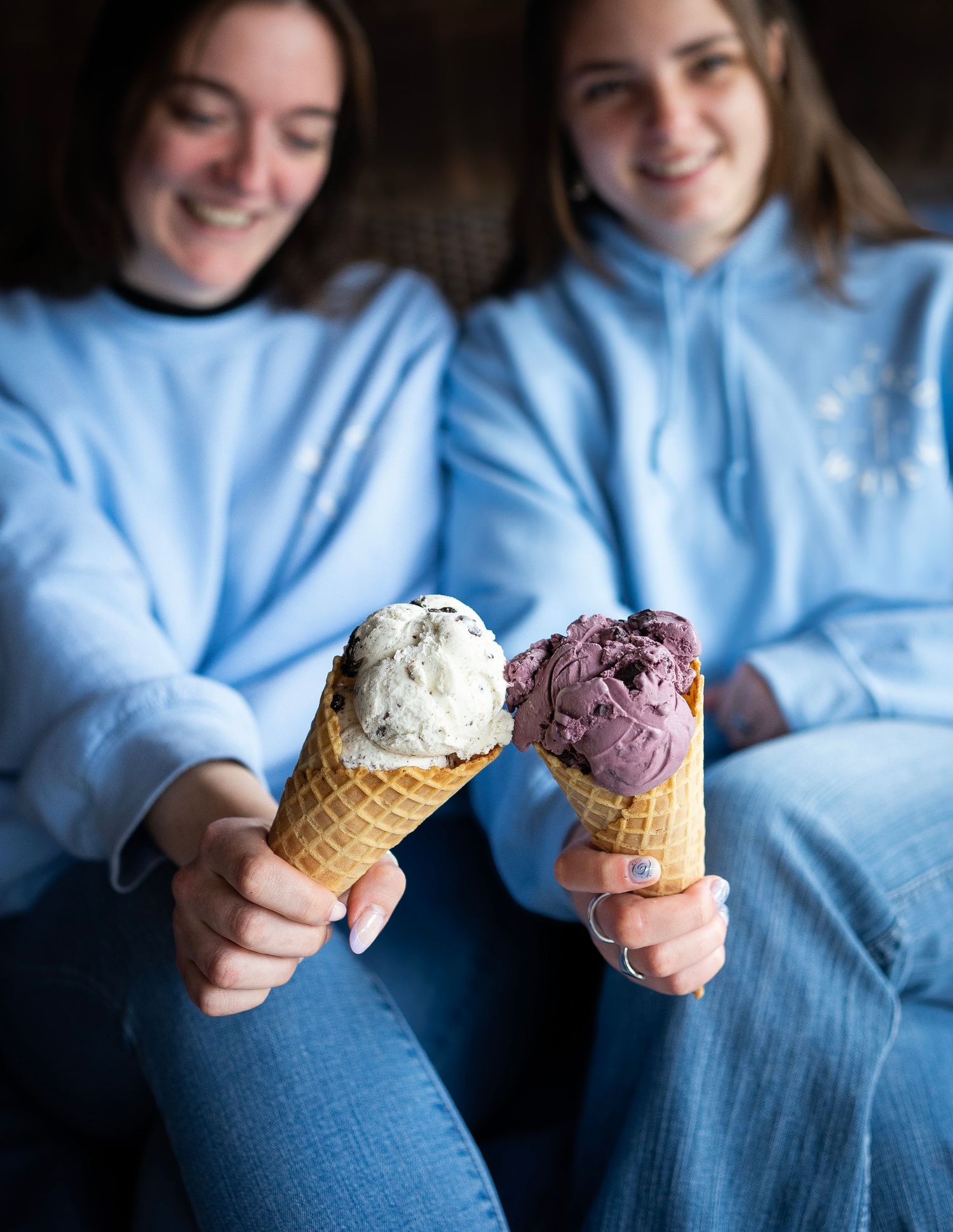 Two people wearing light blue sweatshirts hold ice cream cones with different flavors, one white and one purple. They are smiling.