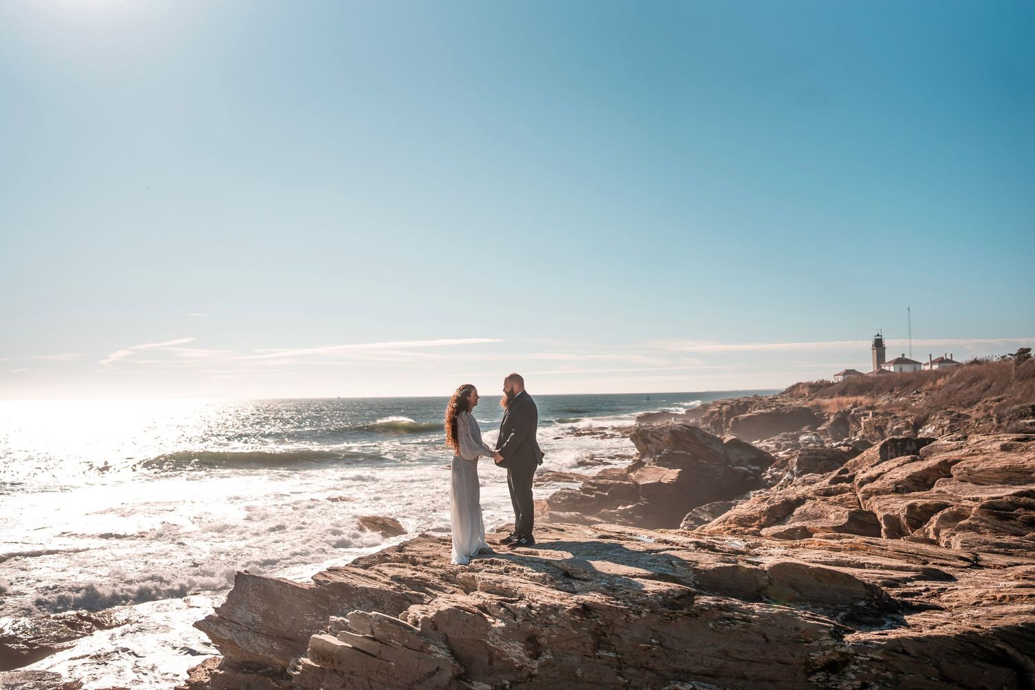 A couple stands facing each other on rocky cliffs by the ocean, holding hands. The sun shines brightly in the clear sky, and a lighthouse.