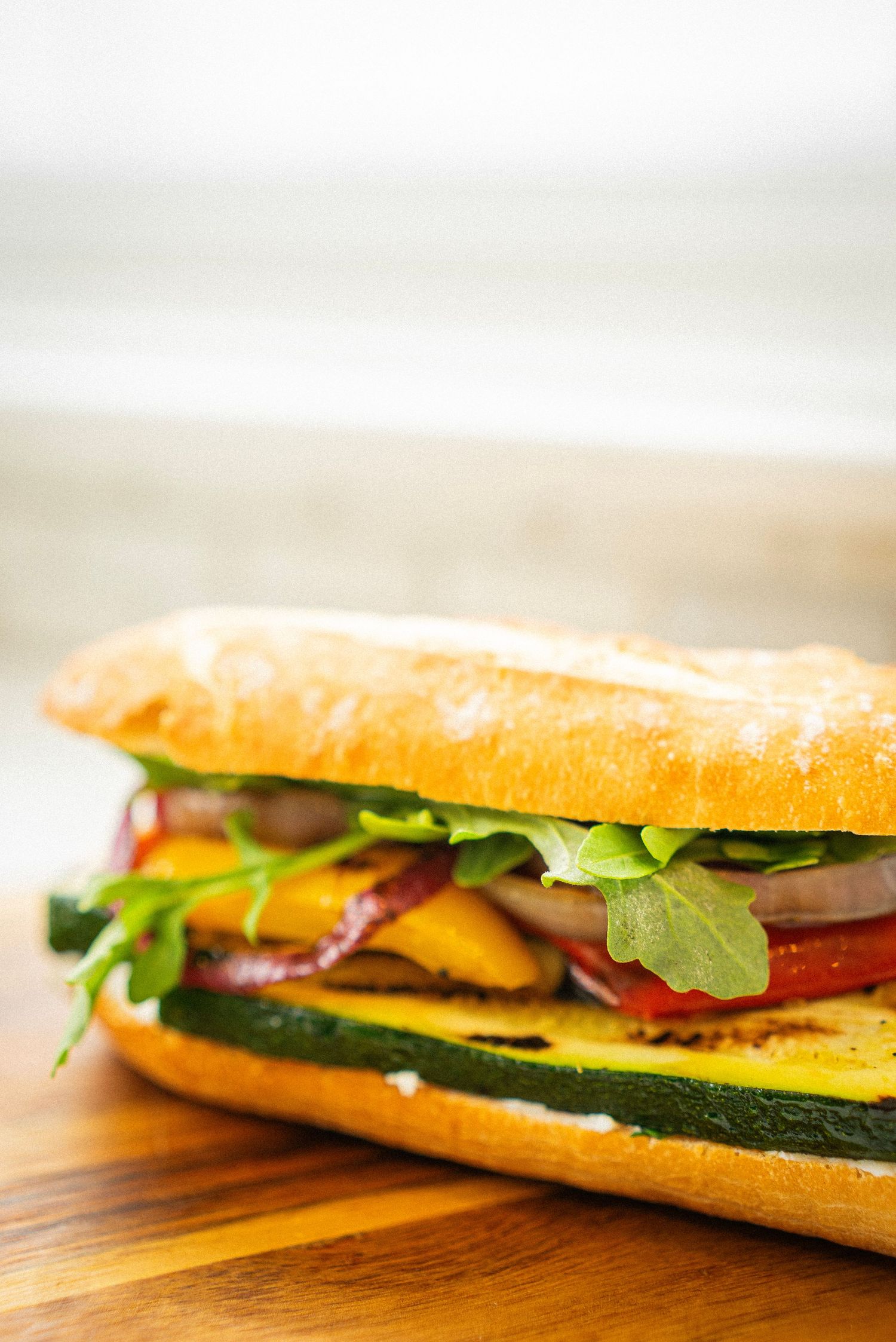 baguette sandwich filled with sliced zucchini, yellow peppers, tomatoes, onions, and arugula sits on a wooden cutting board in natural light