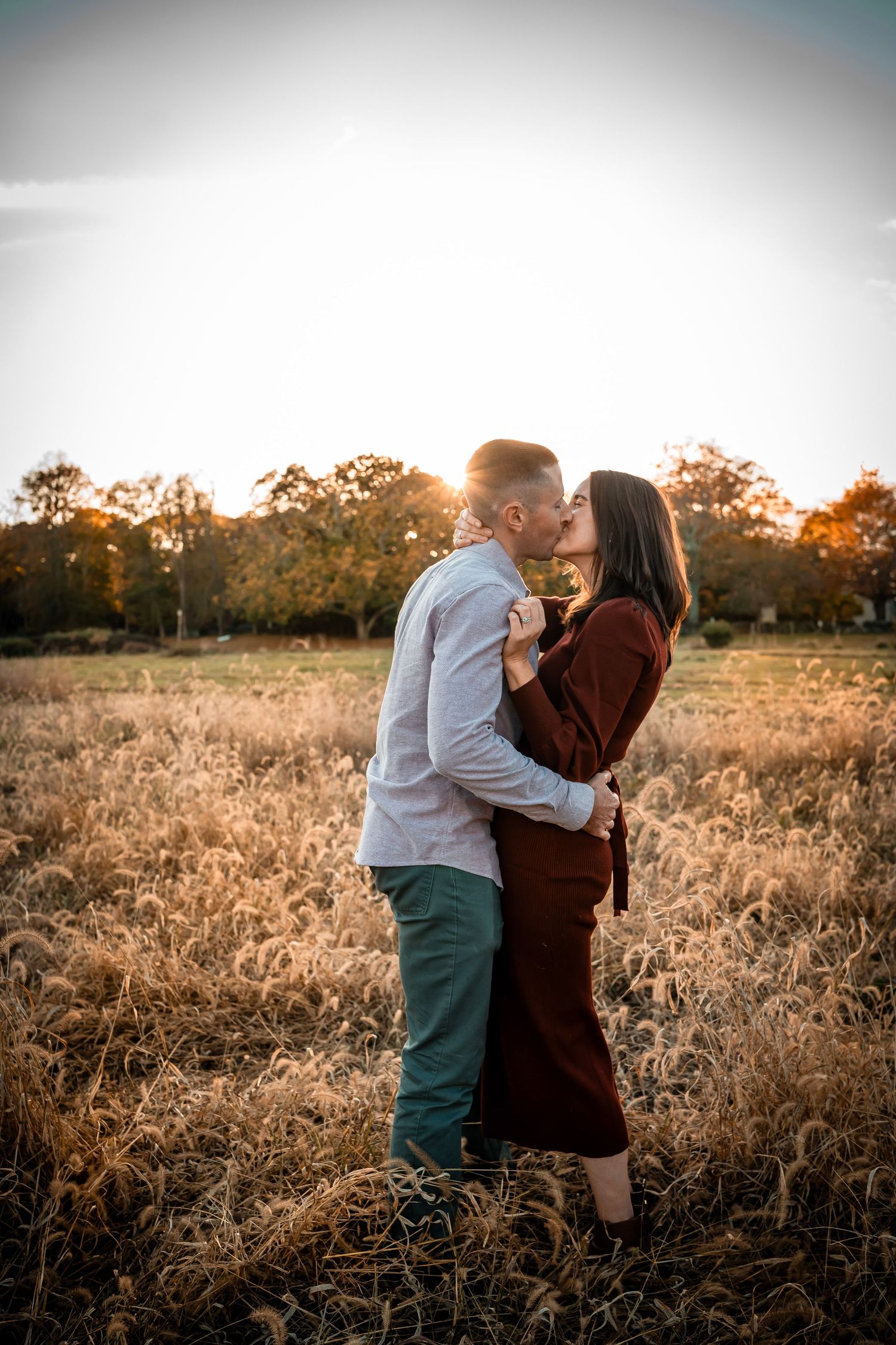 A couple kissing one another in a field during golden hour. The couple dressed in formal attire stands close and are in love.