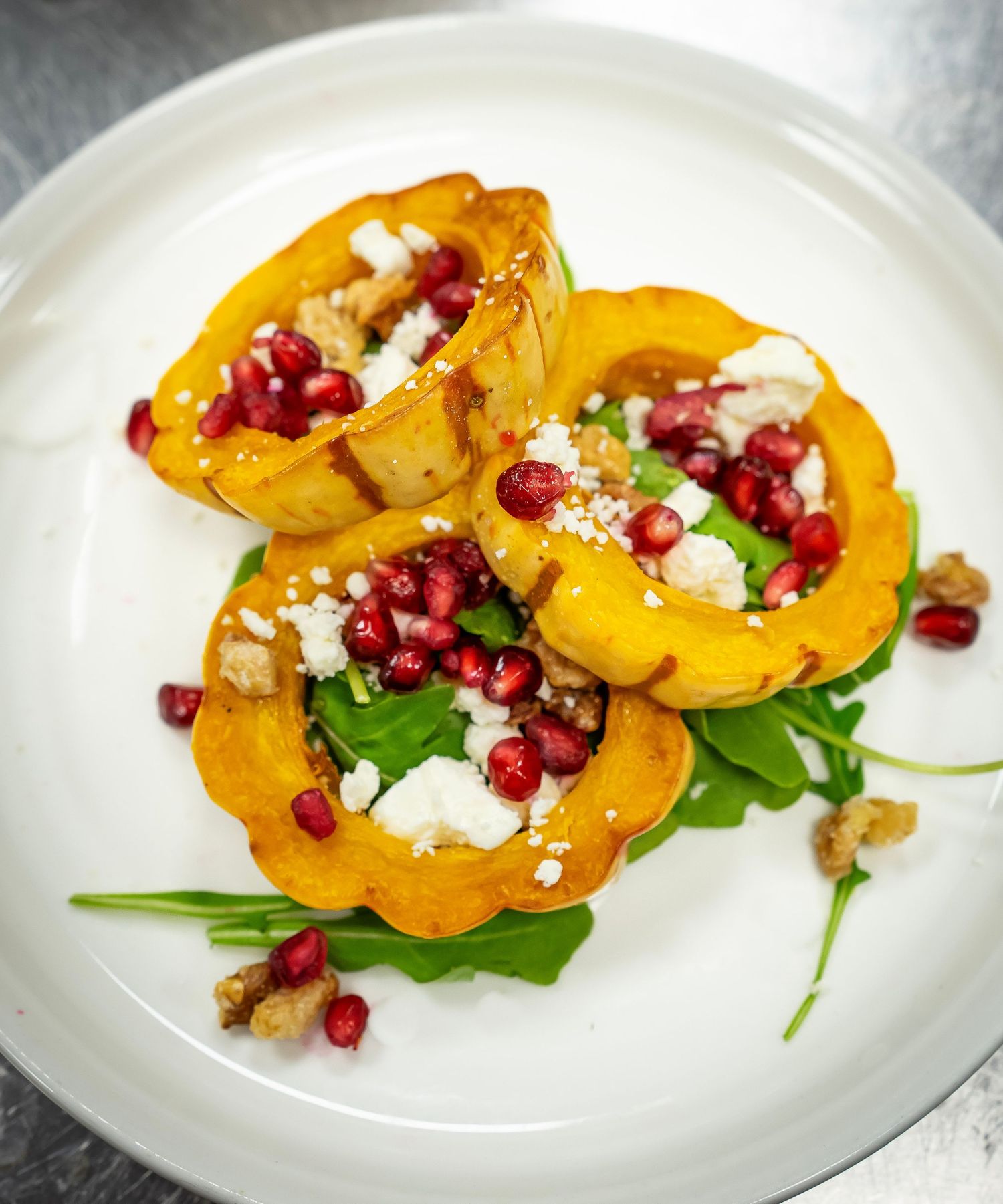 A white plate with roasted acorn squash rings filled with pomegranate seeds, walnuts, feta cheese, and greens. The toppings add rich color