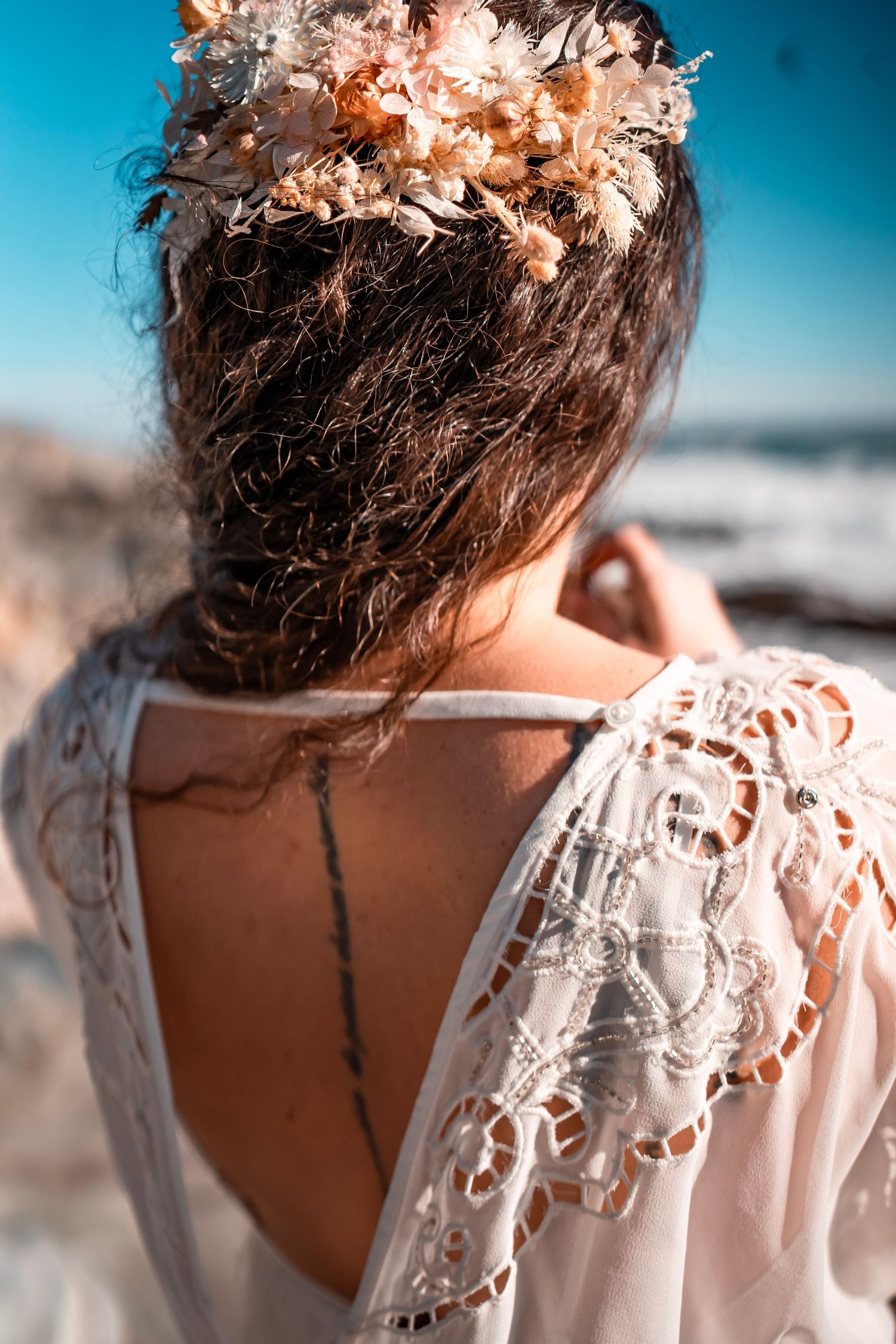 An up-close photograph of a bohemian bride with florals in her hair, white dress, and tattoo down her back. Sun shines bright and  ocean.