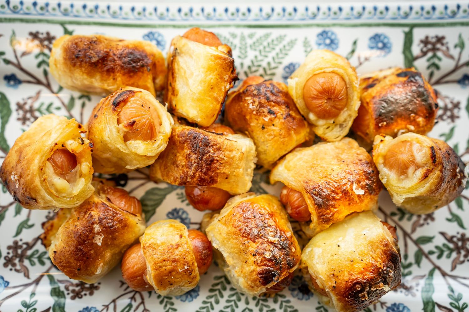 A decorative platter holds several golden-brown pigs in a blanket, with sausages wrapped in flaky pastry. It showcases their crispy texture.