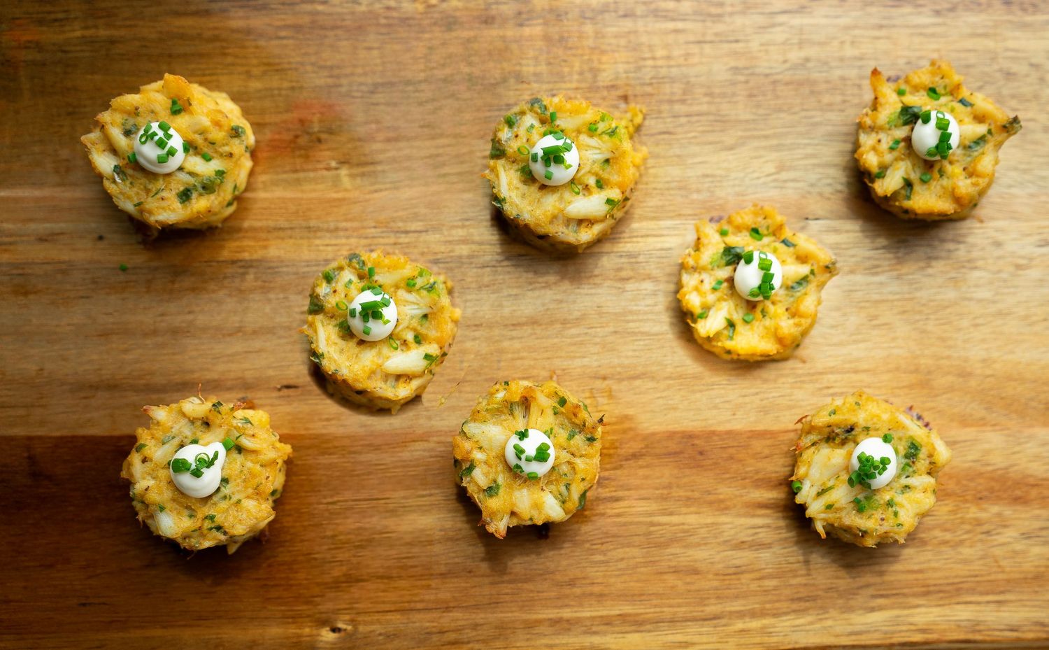 Mini crab cakes topped with a dollop of sauce and garnished with green herbs are artfully arranged on a wooden surface, perfect for catering