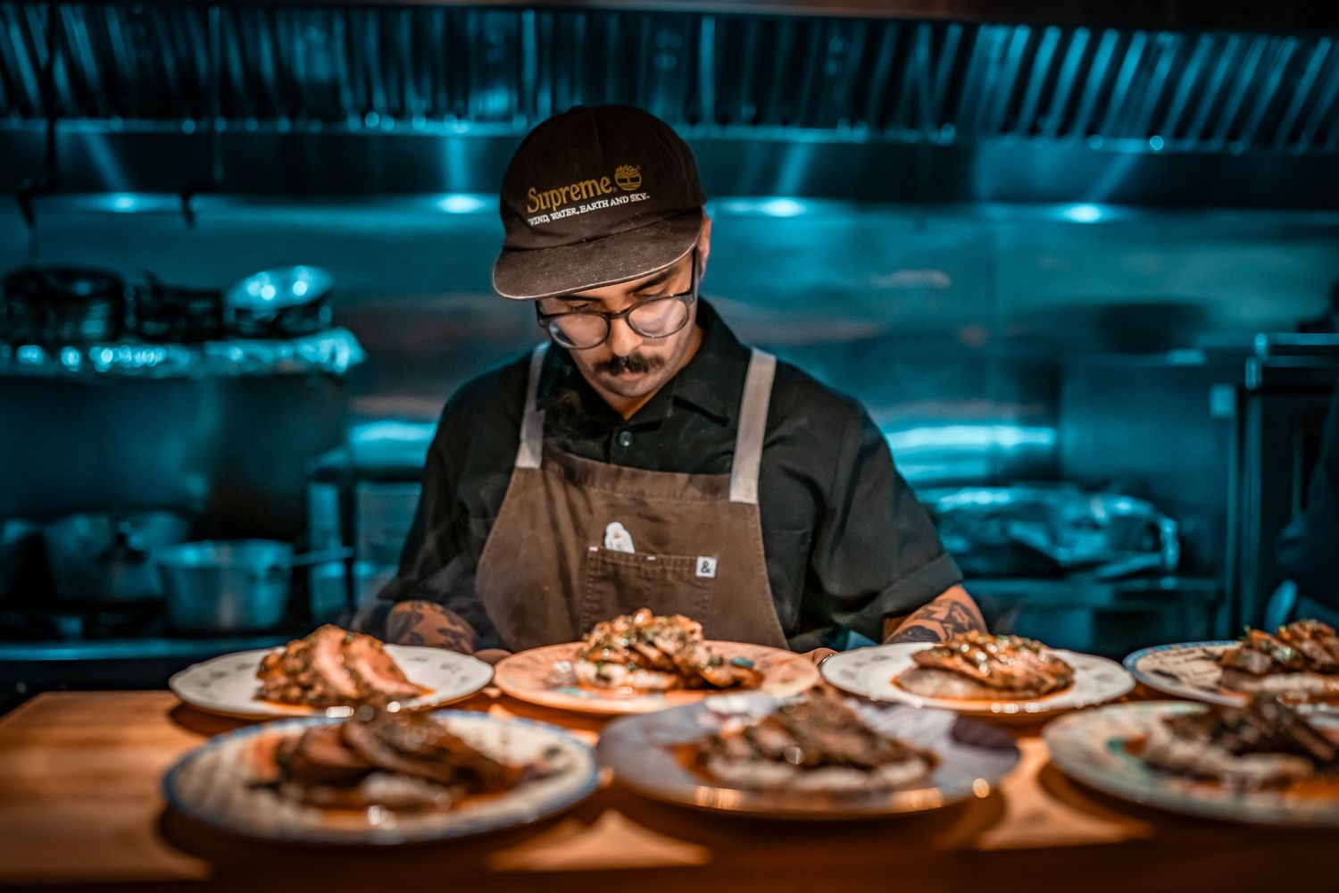 A chef stands behind a counter, plating dishes in a dimly lit kitchen. Plates of food are arranged in front of him, showcasing various meat.