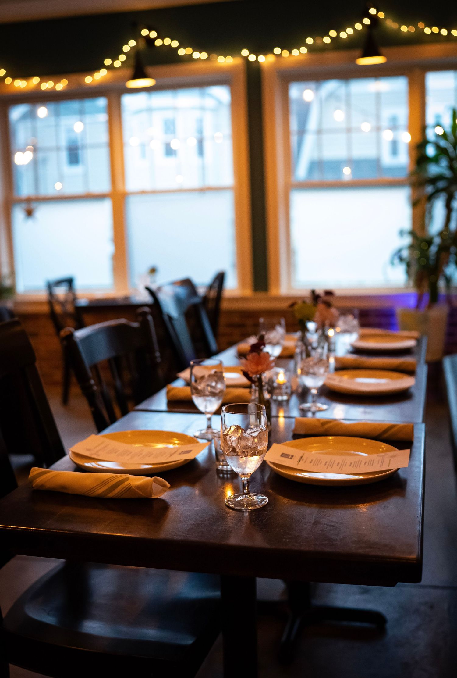 A dimly lit restaurant features a table set for several people. Plates, glasses, and napkins neatly arranged. String light. Cozy atmosphere