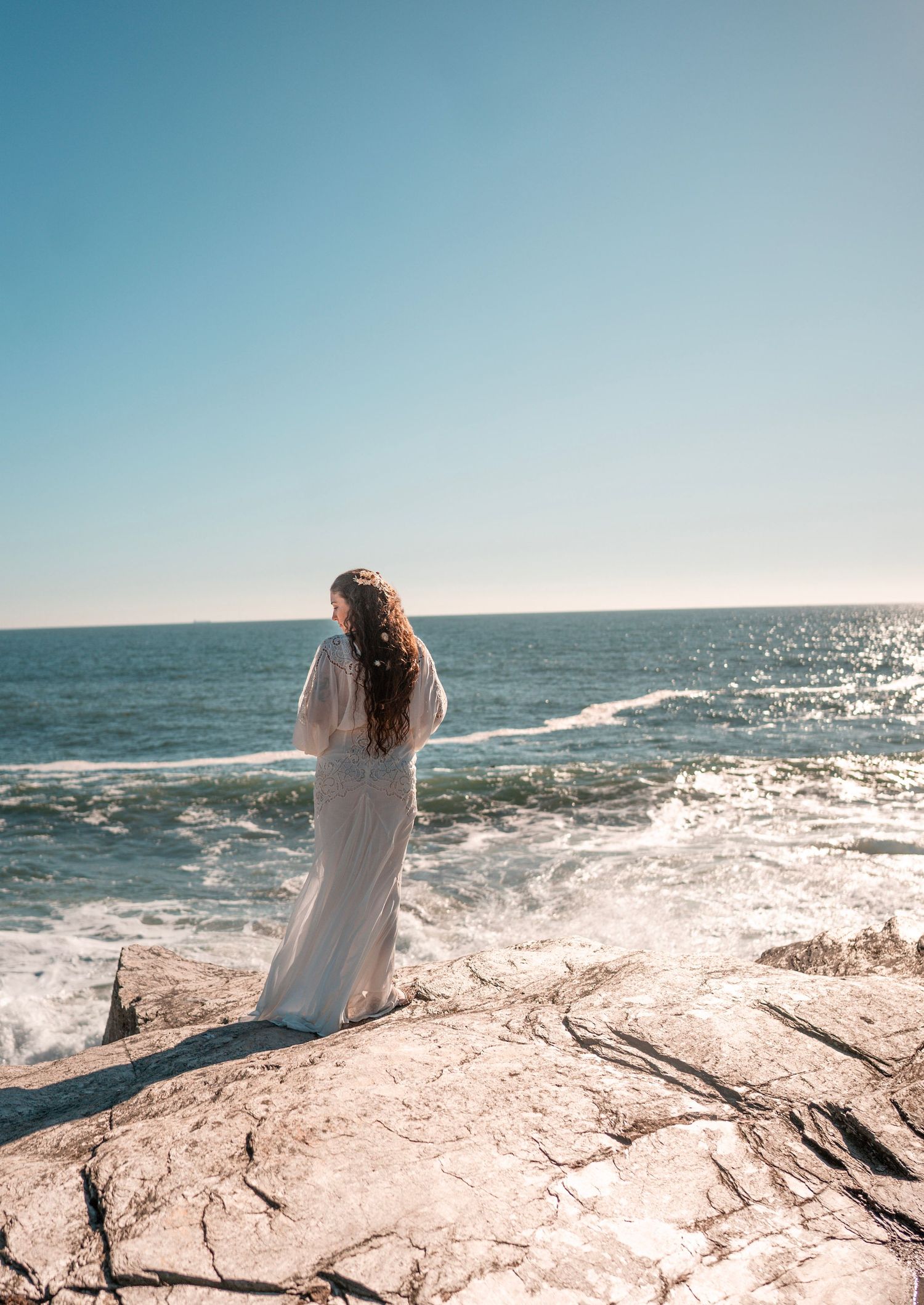 An up-close photograph of a bohemian bride with florals in her hair, she stand on the rocks as the ocean and waves crash behind her.