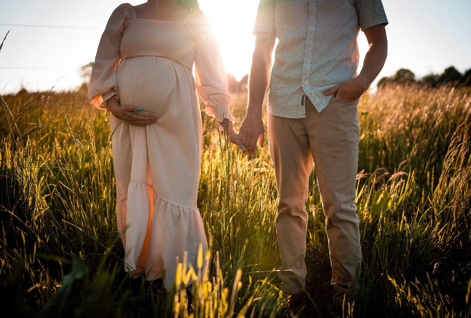 A pregnant woman in a light dress and a man in a shirt and beige pants hold hands in a grassy field, with sunlight softly illuminating them.