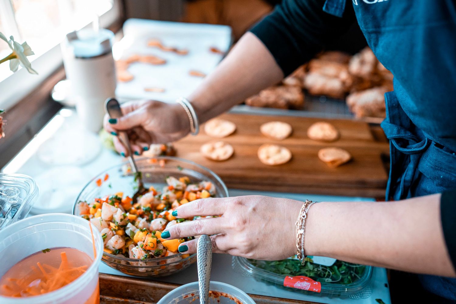 A person prepares ceviche in a glass bowl, adding fresh ingredients with a spoon. Nearby, crackers and herbs are placed on a wooden cutting