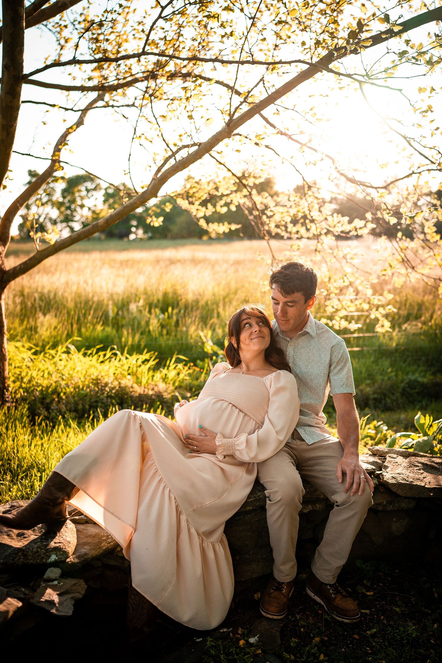 The woman is pregnant, wearing a flowing dress. She is leaning on the man looking at each other. Sunlight filters through the leaves.