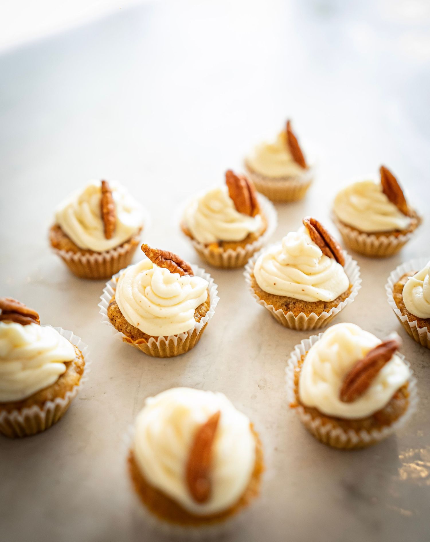 Mini cupcakes topped with swirls of creamy frosting and pecan halves are arranged on a white marble surface. The cupcakes are in small paper
