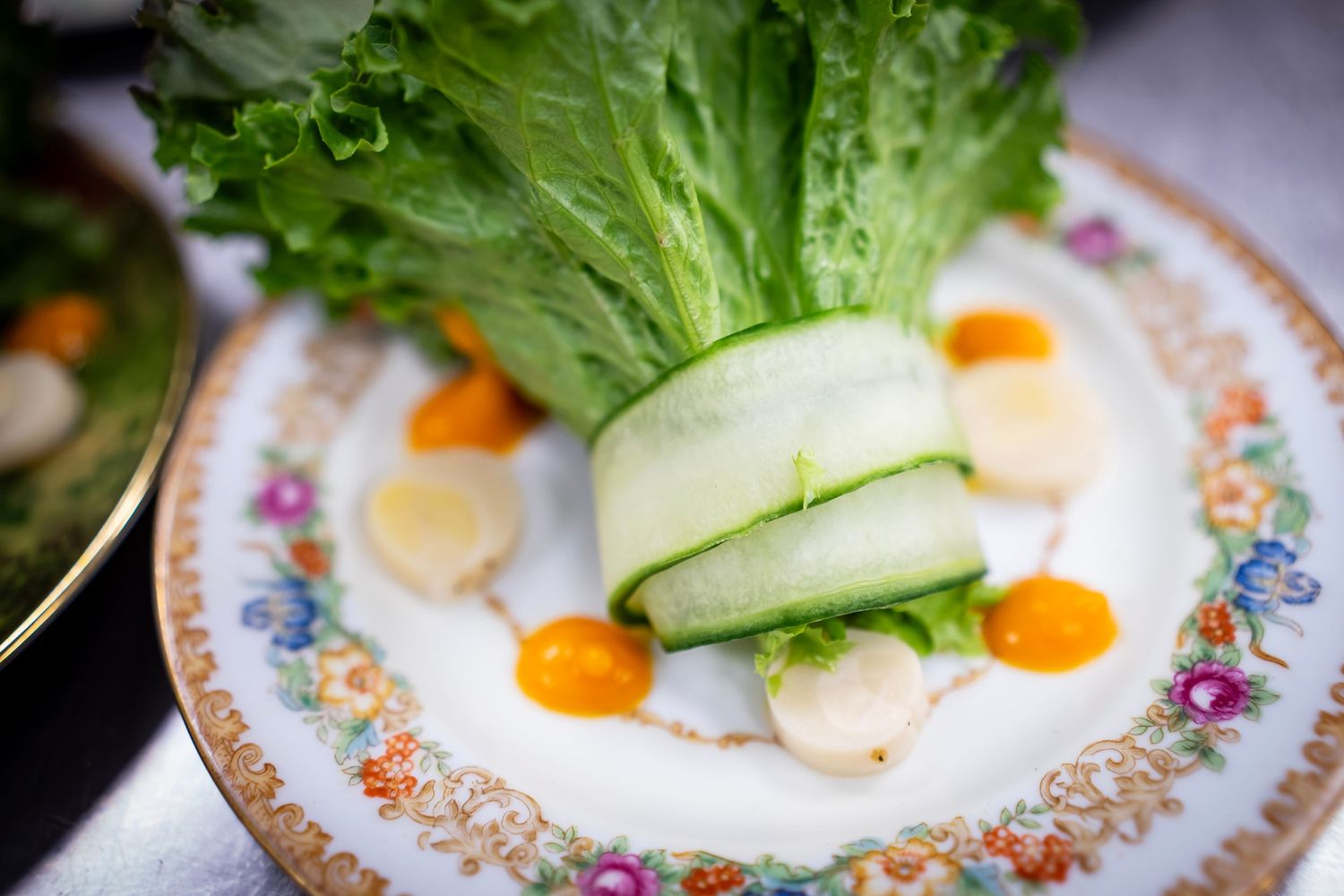 A photo of a decorative dish features a bundle of green lettuce wrapped with cucumber slices, placed on a white plate with a floral pattern.