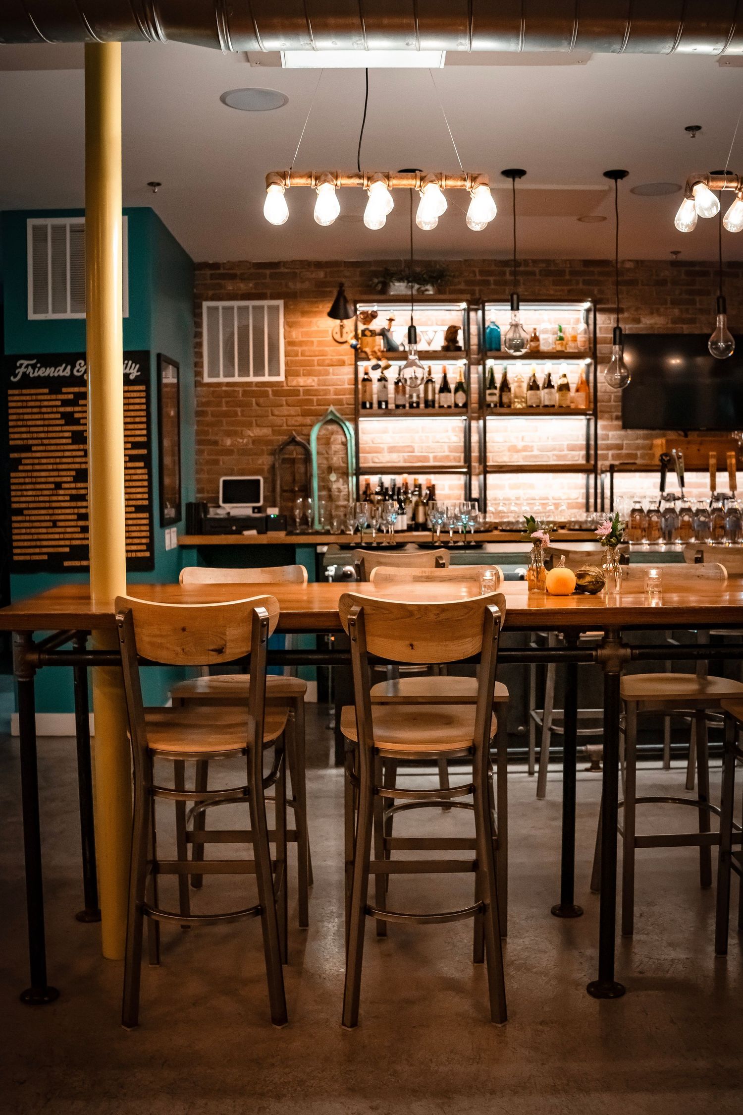 A cozy bar setting with wooden chairs lined up along a metal counter. Warm hanging lightbulbs illuminate the space. A brick wall and bar.