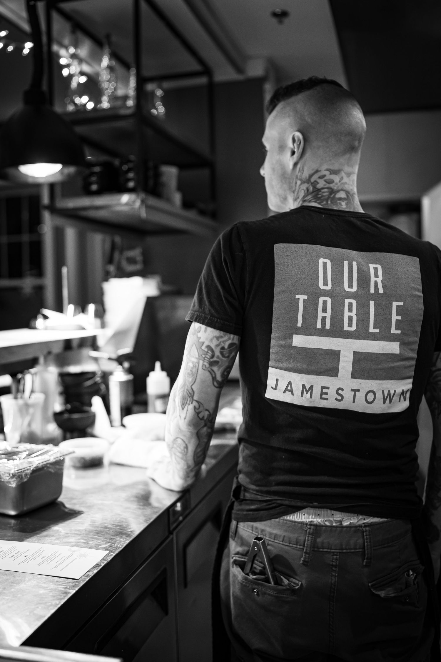A person with tattoos wearing a T-shirt with OUR TABLE JAMESTOWN on the back stands at kitchen counter. The setting is a restaurant kitchen