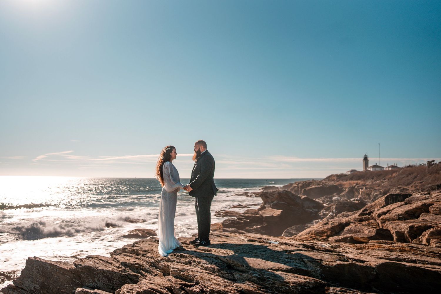 A couple stands facing each other on rocky cliffs by the ocean, holding hands. The sun shines brightly in the clear sky, and a lighthouse.
