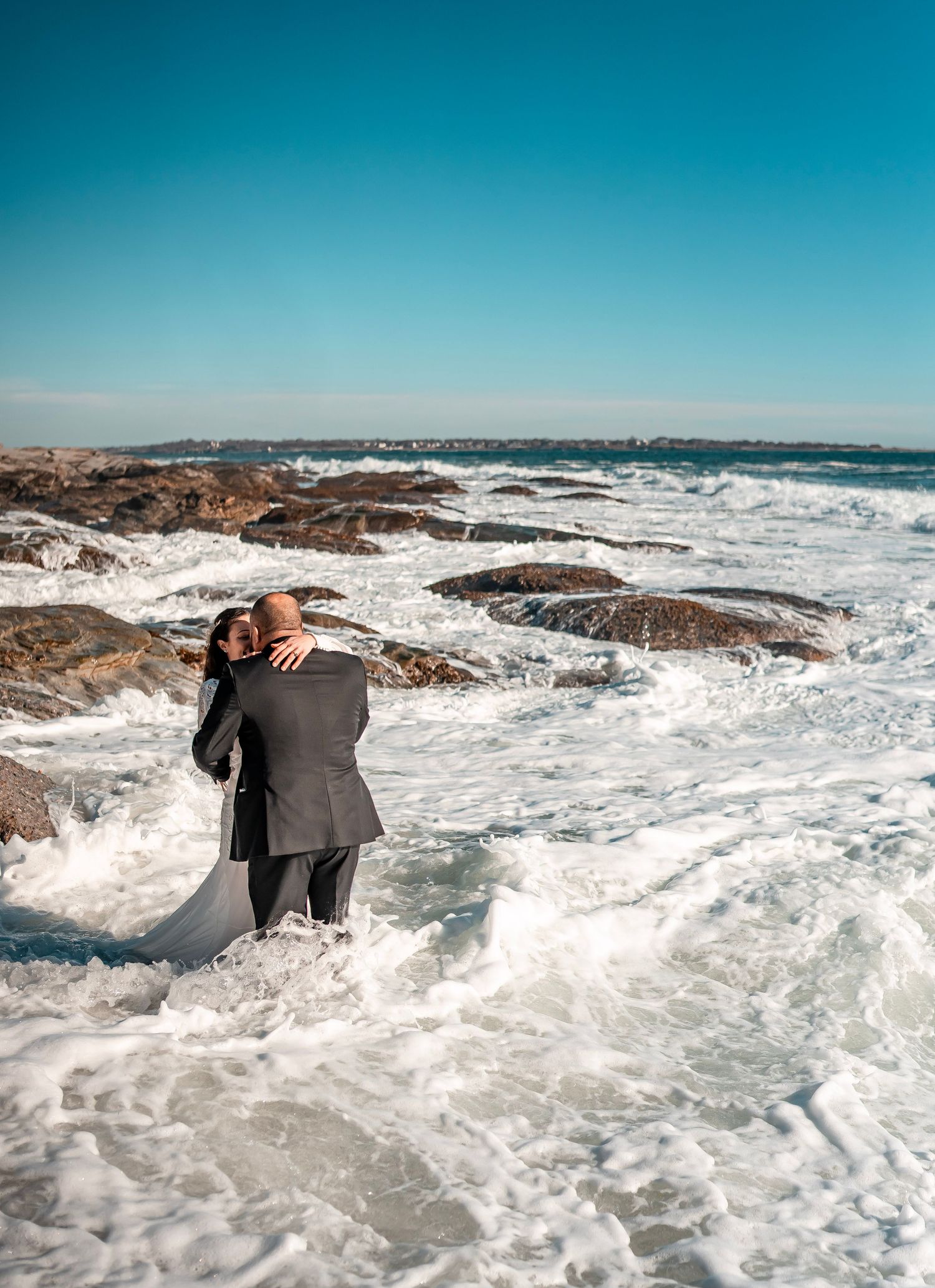 A bride and groom is photographed kissing in the ocean with the waves crashing around them as they kiss.