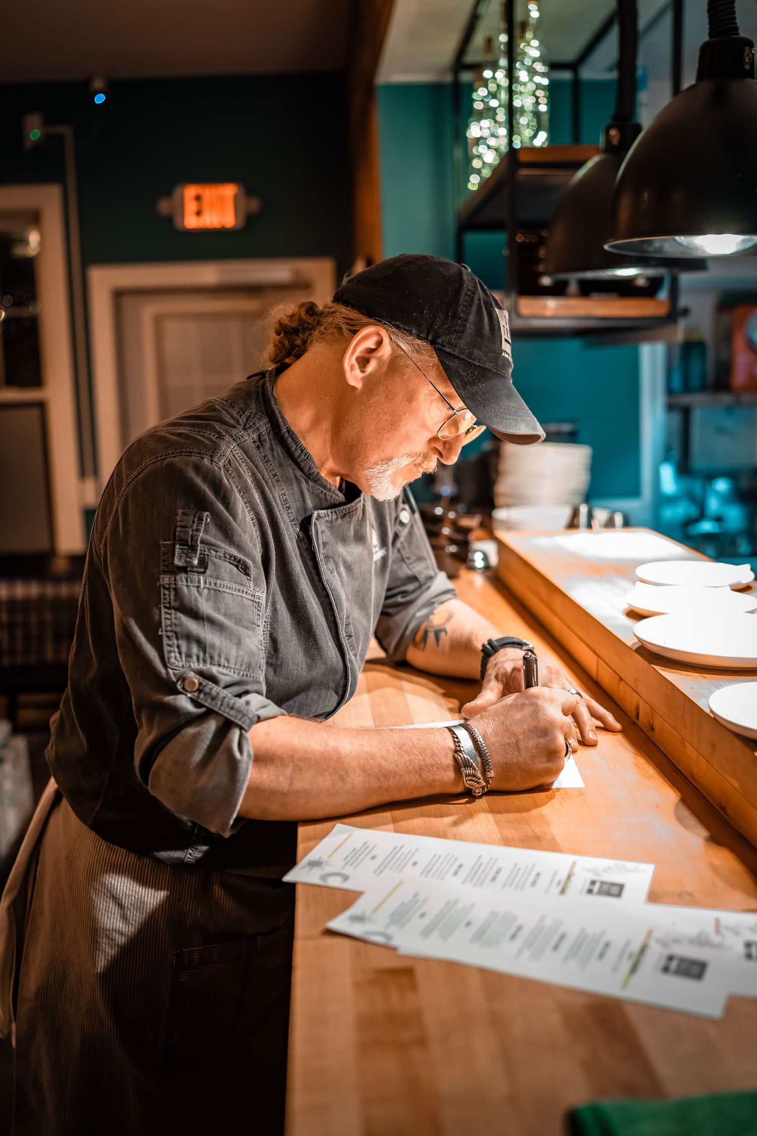 Chef in a baseball cap and glasses writing on papers at a wooden counter in a restaurant kitchen. Plates are arranged under hanging lights.
