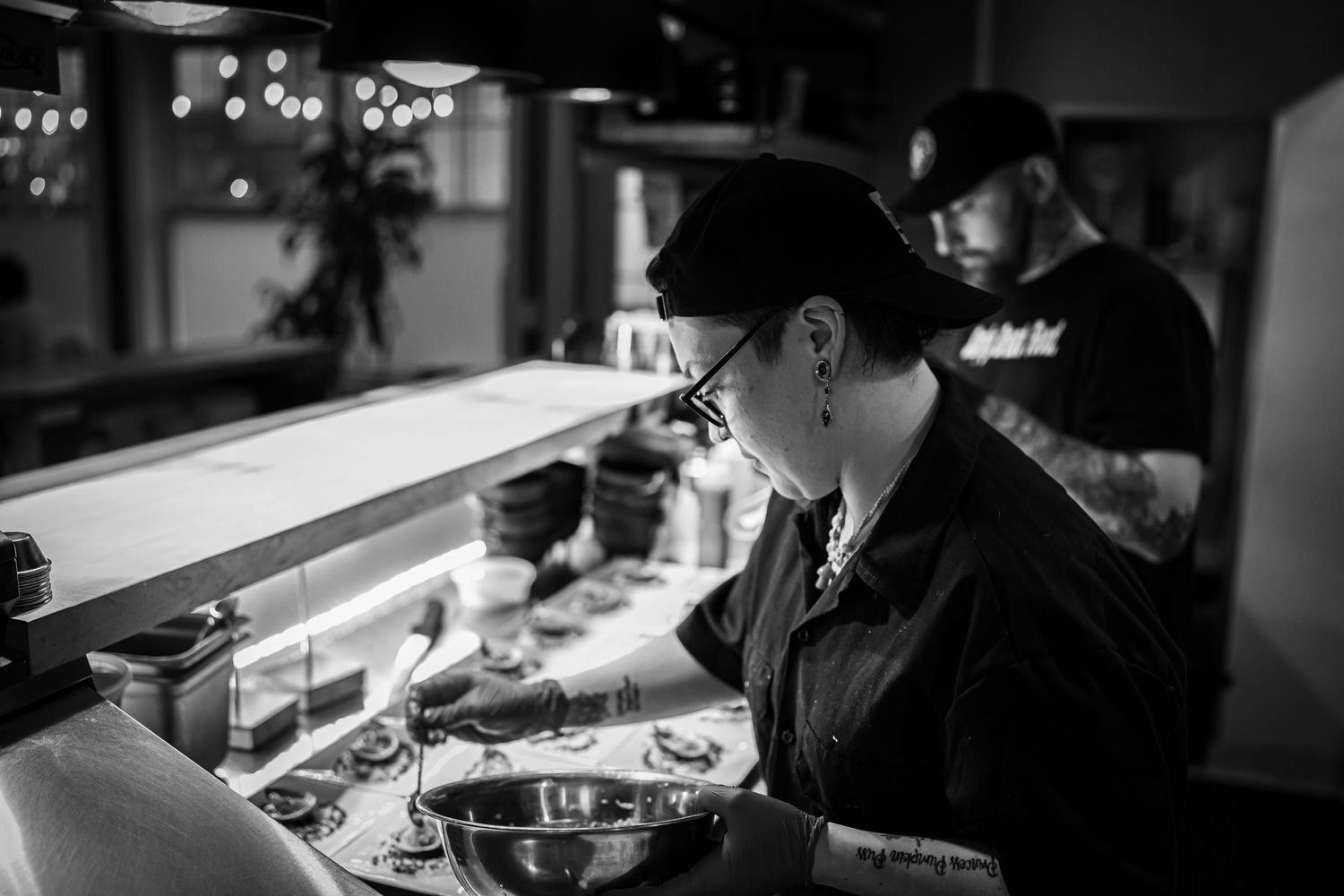 A chef wearing a cap and glasses is focusing on plating dishes in a restaurant kitchen. Another chef works in the background. Kitchen setup.