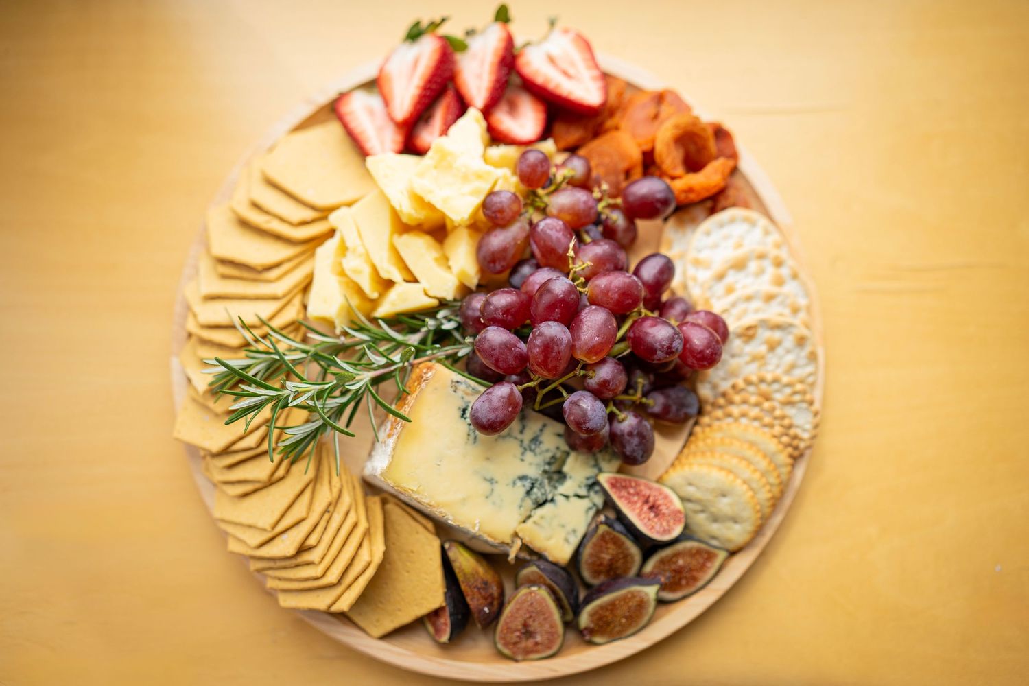 A gourmet cheese platter featuring blue cheese, crackers, figs, red grapes, and assorted fruits displayed artfully on a round wooden board.