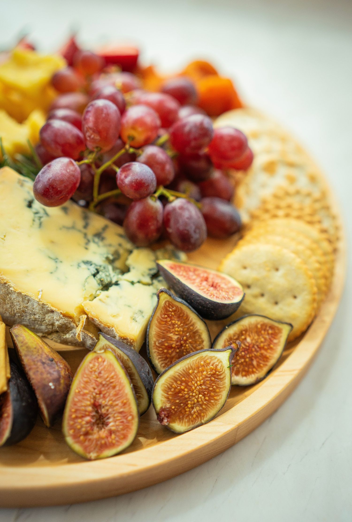 A gourmet cheese platter featuring blue cheese, crackers, figs, red grapes, and assorted fruits displayed artfully on a round wooden board.
