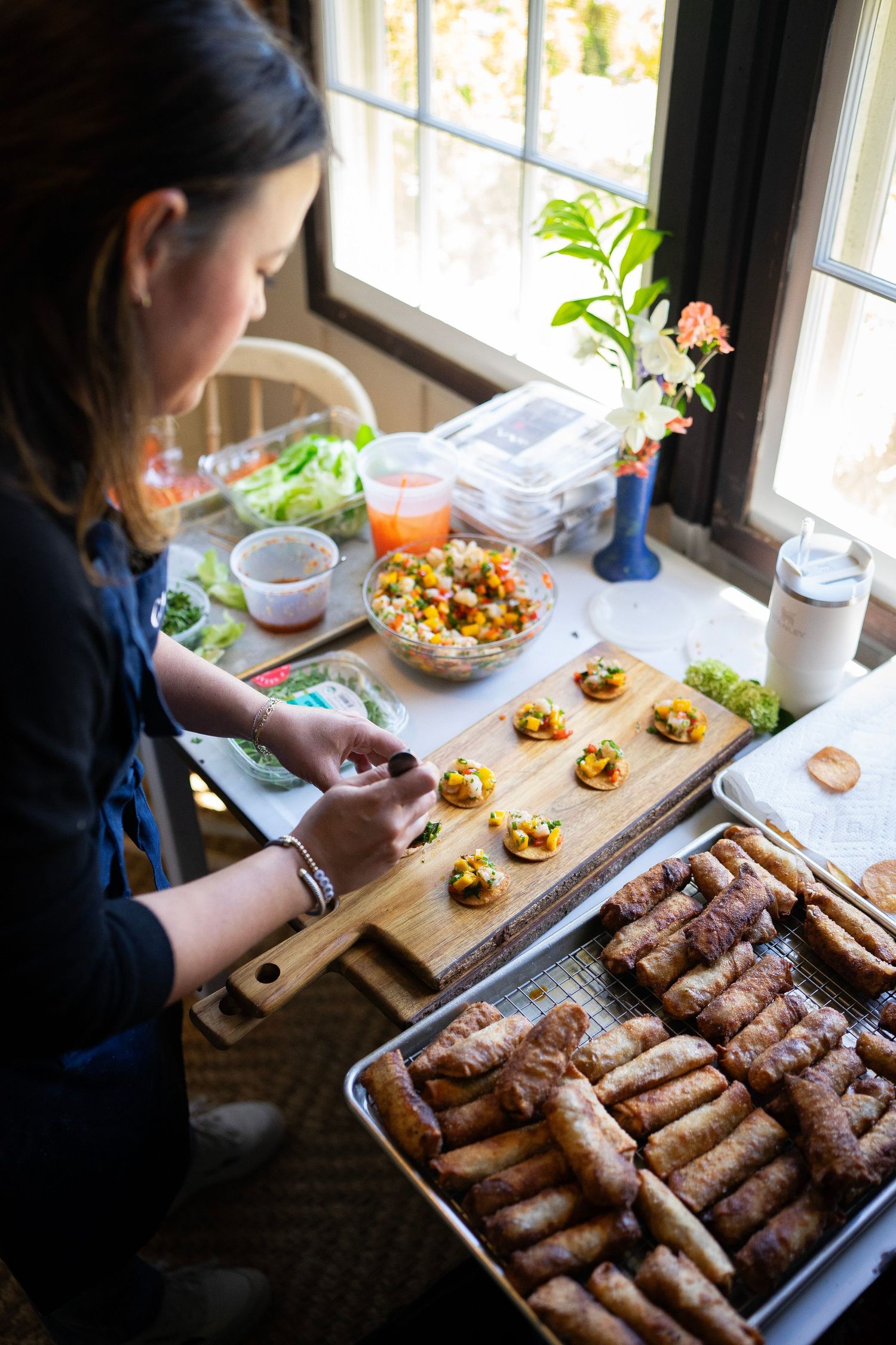 A person in a black shirt is preparing food on a wooden board next to a window. Various dishes, including spring rolls and colorful salads.
