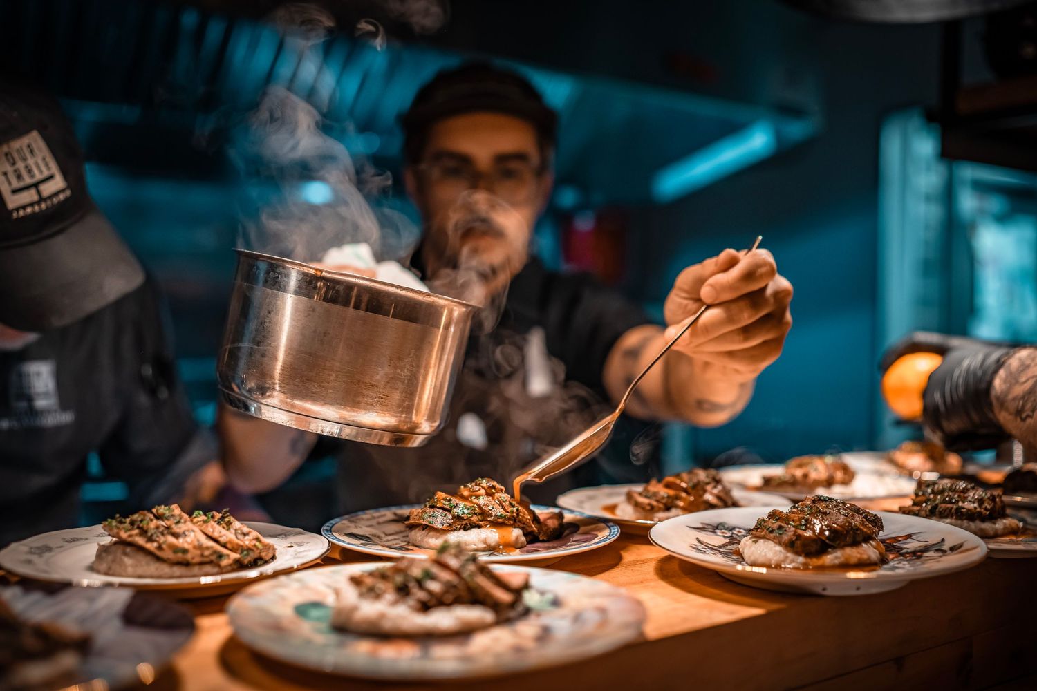A chef stands behind a counter, plating dishes in a dimly lit kitchen. Plates of food are arranged in front of him, showcasing various meat.