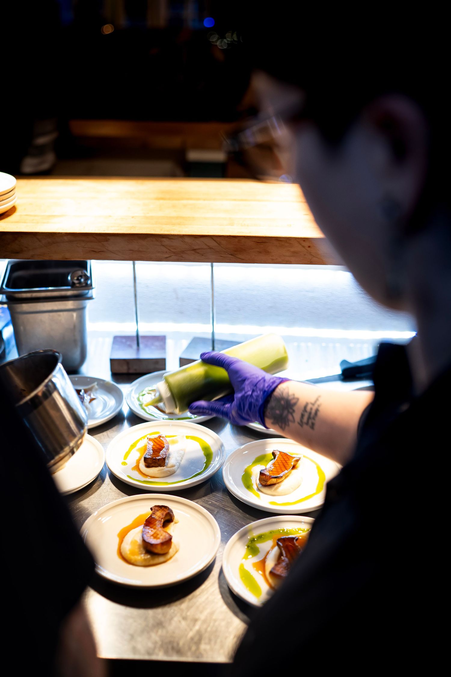 A chef in a kitchen prepares plates of gourmet food, adding sauce from a squeeze bottle. The dishes feature seared meat, a dollop of sauce.
