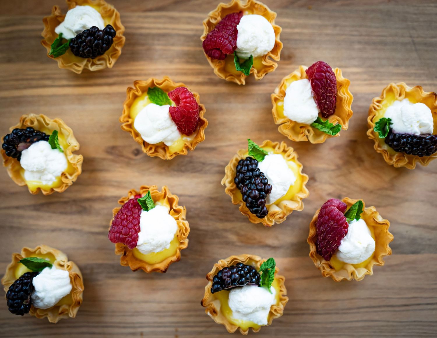 A photograph of mini pastry cups topped with cream, raspberries, blackberries, and mint leaves are arranged on a wooden surface.