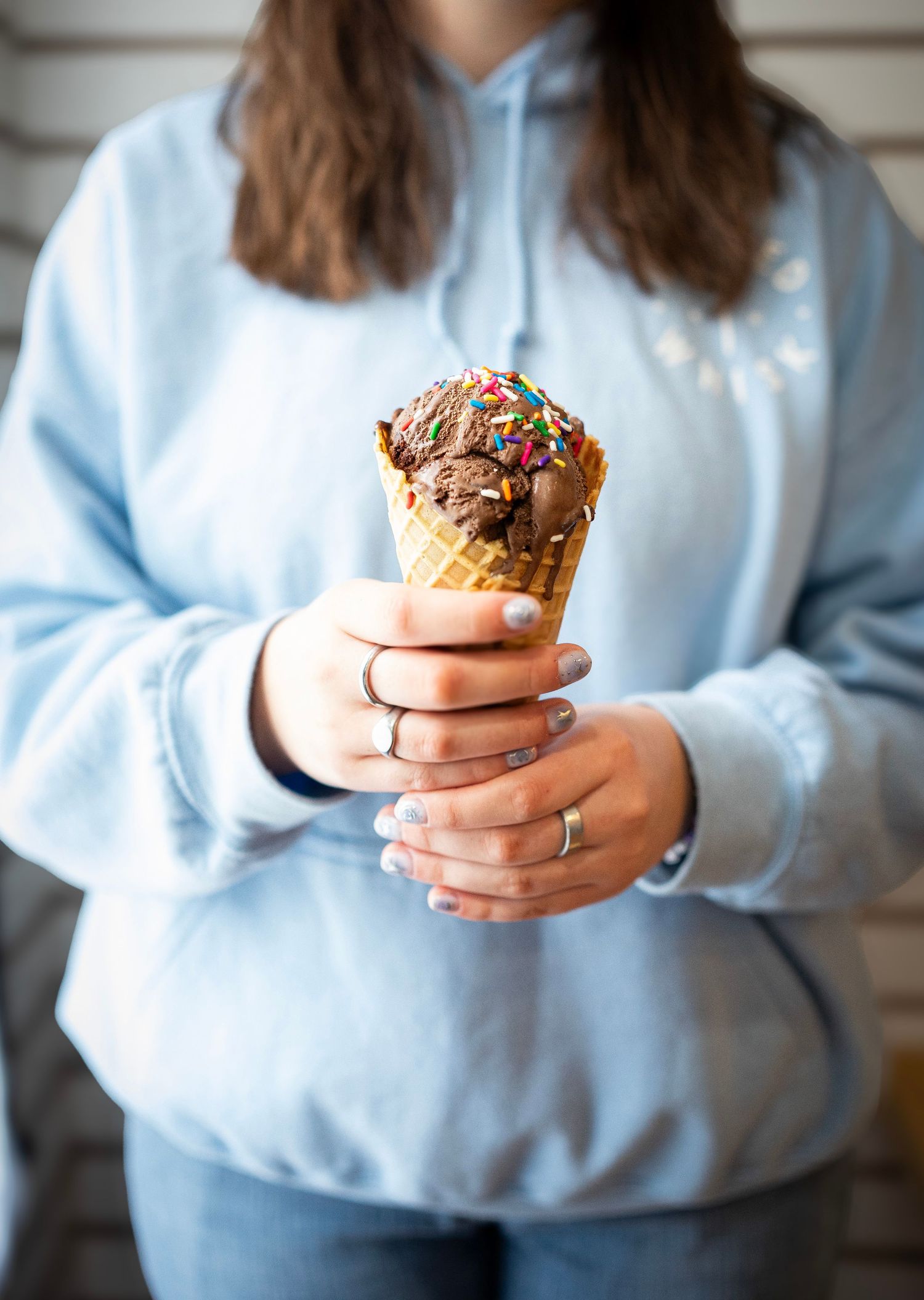 A person wearing a light blue sweatshirt holds a waffle cone with a scoop of pink ice cream topped with colorful sprinkles.