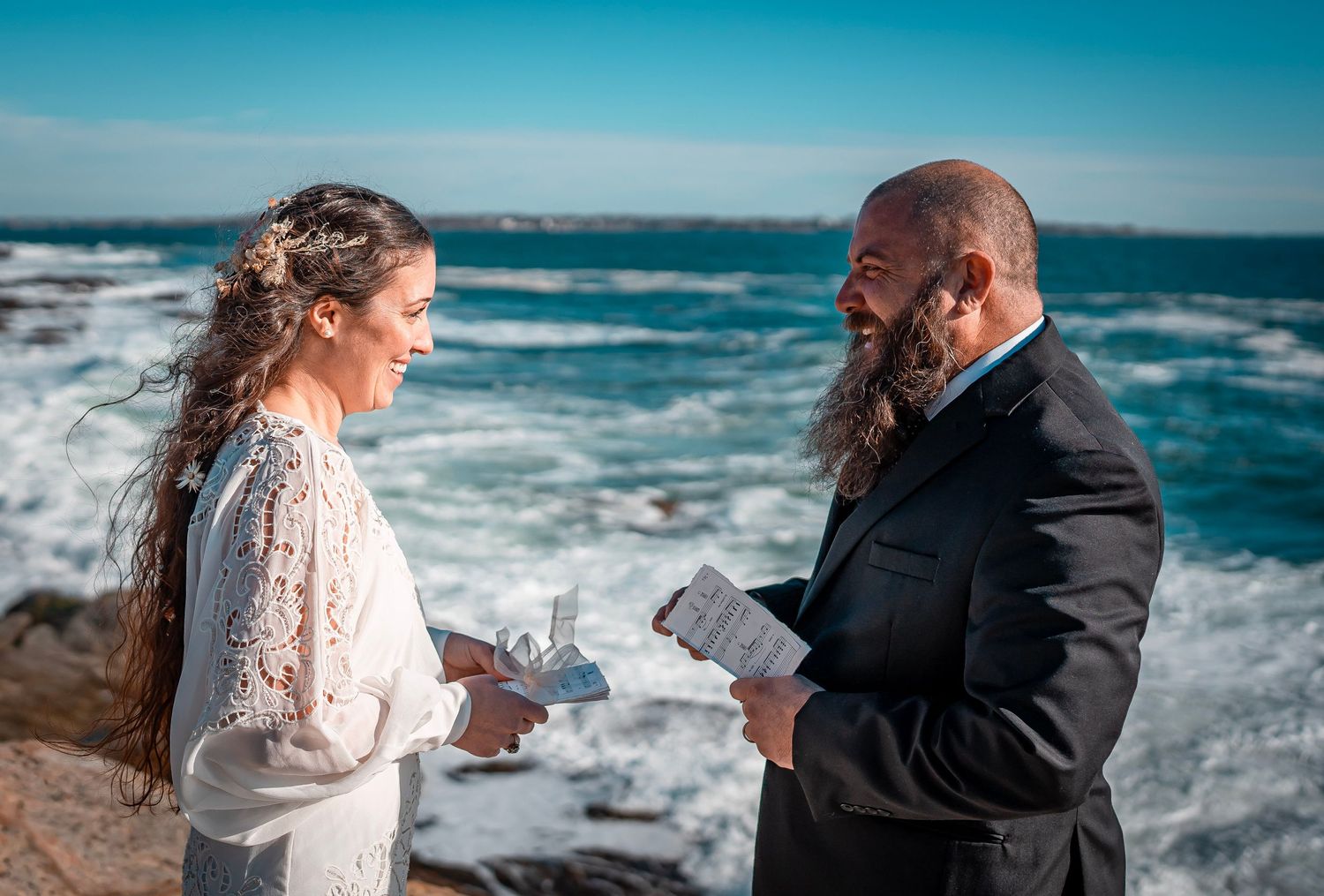 A couple stands facing each other on rocky cliffs by the ocean saying their vows. The water is blue and they are renewing their vows.