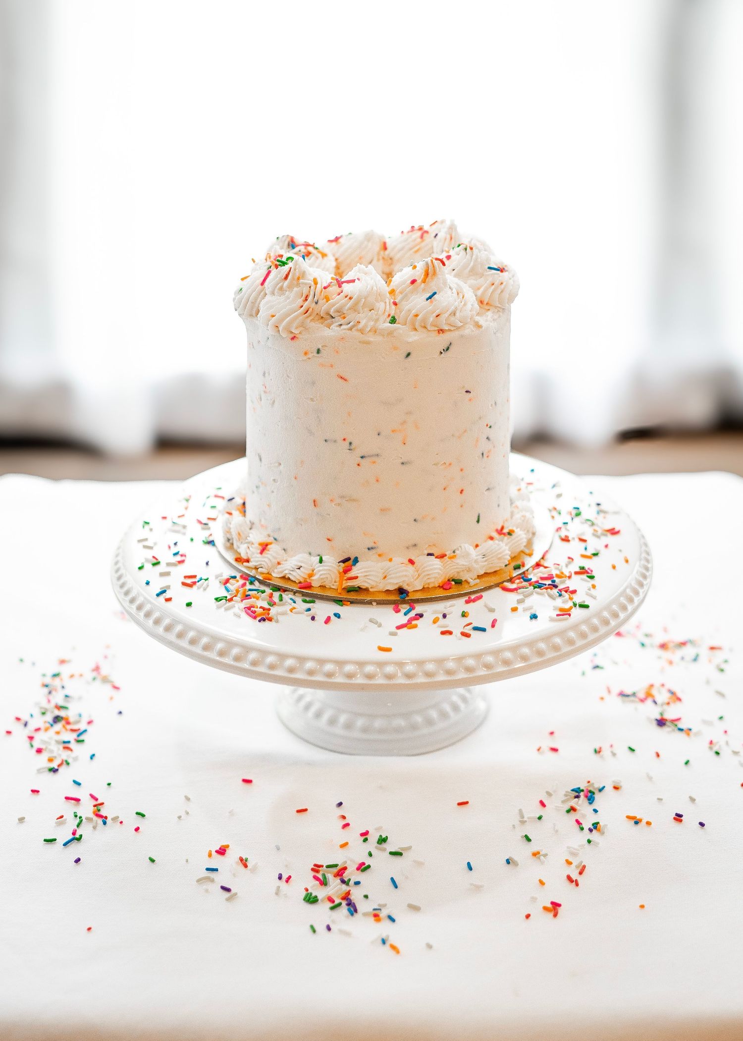 A beautifully photographed cake on a white display tray with sprinkles. A funfetti-style cake made by Wicked Whisk Bakery and white frosting