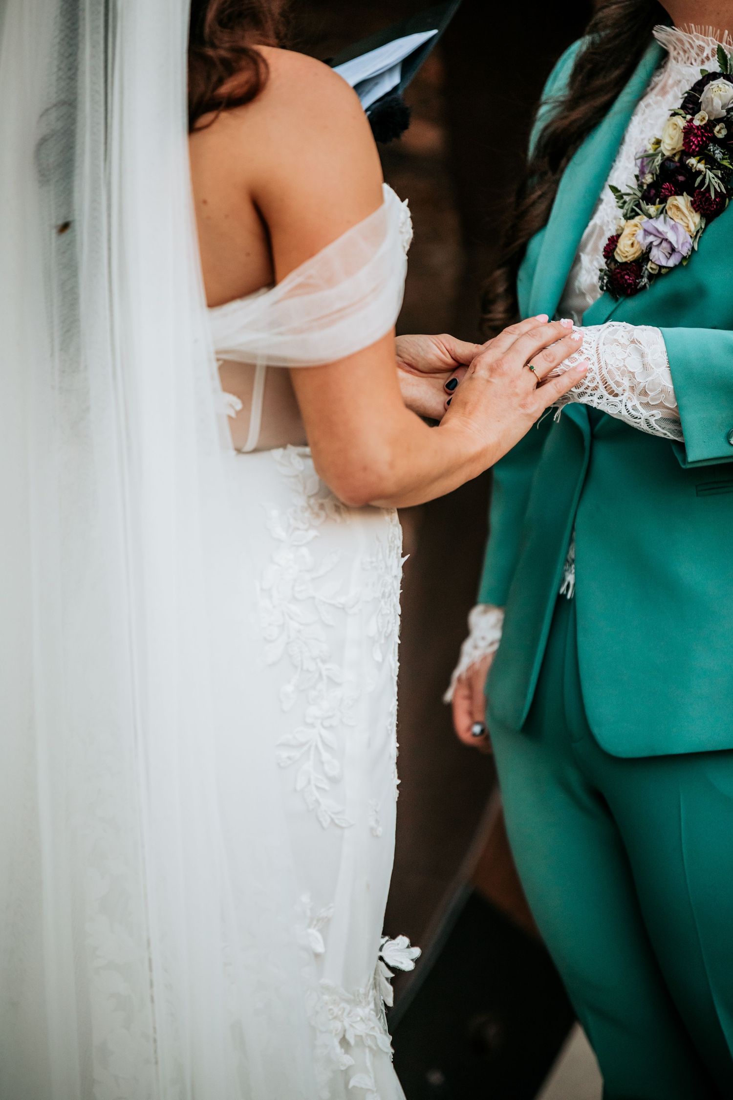 Close-up of two brides holding hands, one in white lace gown and one in teal suit with boutonniere.