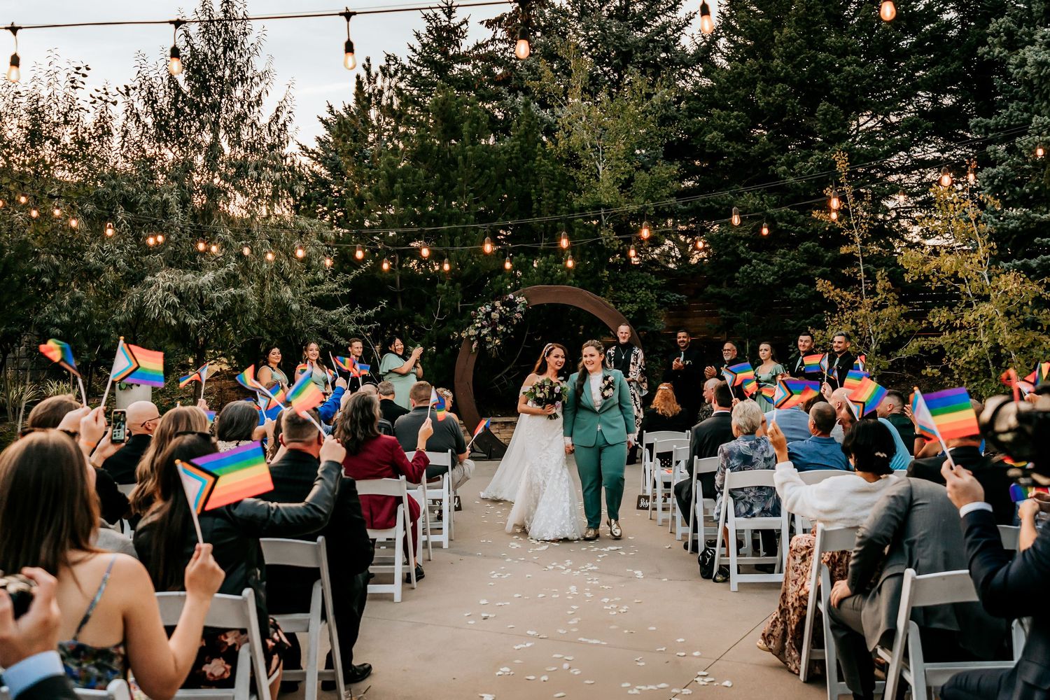 Guests wave pride flags as two brides walk down the aisle at a festive outdoor garden wedding ceremony.