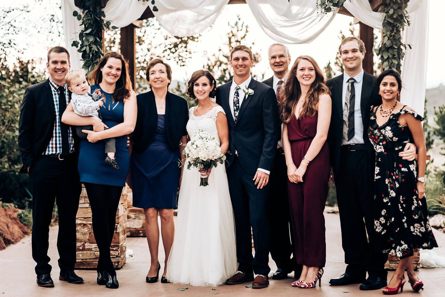 Wedding party gathered under elegant white draped arch with floral decorations.