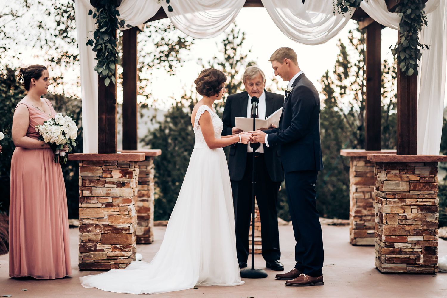 Wedding ceremony under decorated pergola with stone columns and white fabric draping.