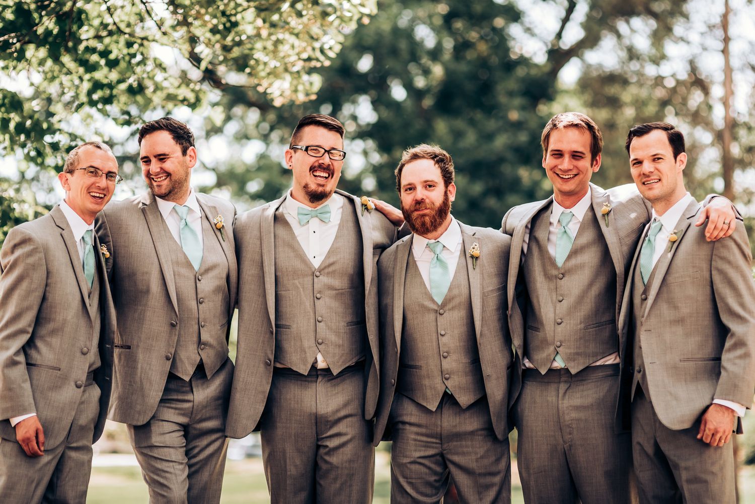 Groomsmen in coordinated gray and blue suits smile together at an outdoor wedding.