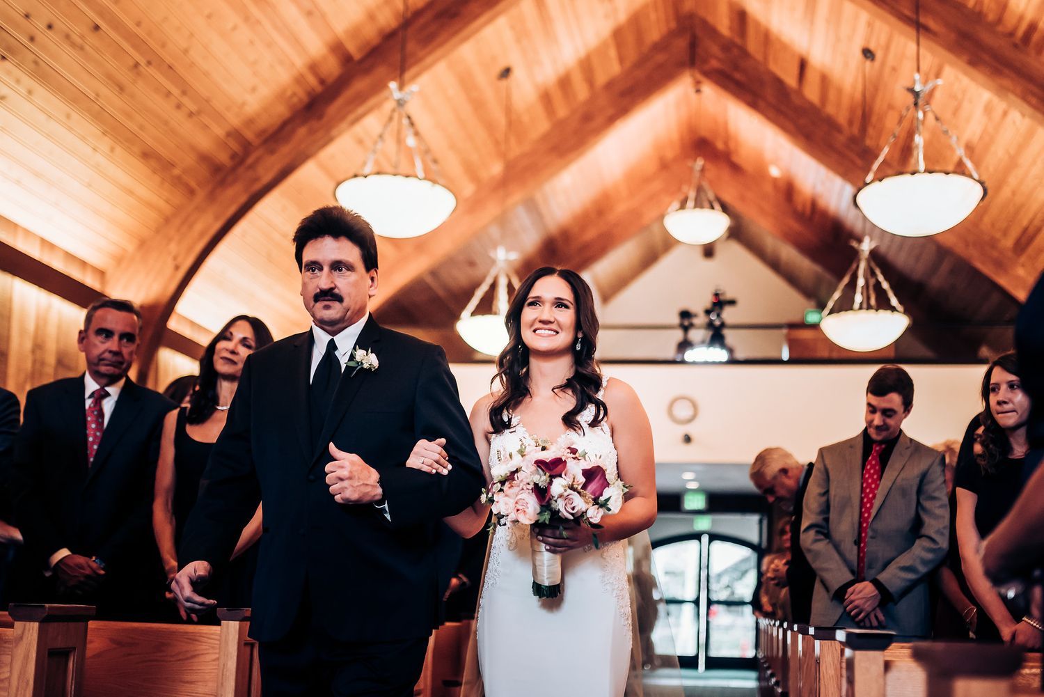A wedding ceremony processional in a wooden-beamed chapel with hanging pendant lights.