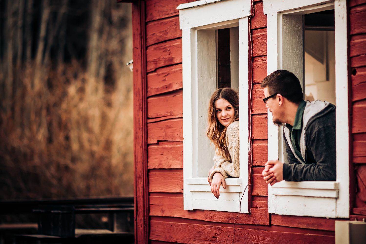 A couple shares laughter while looking through separate windows of a red building.