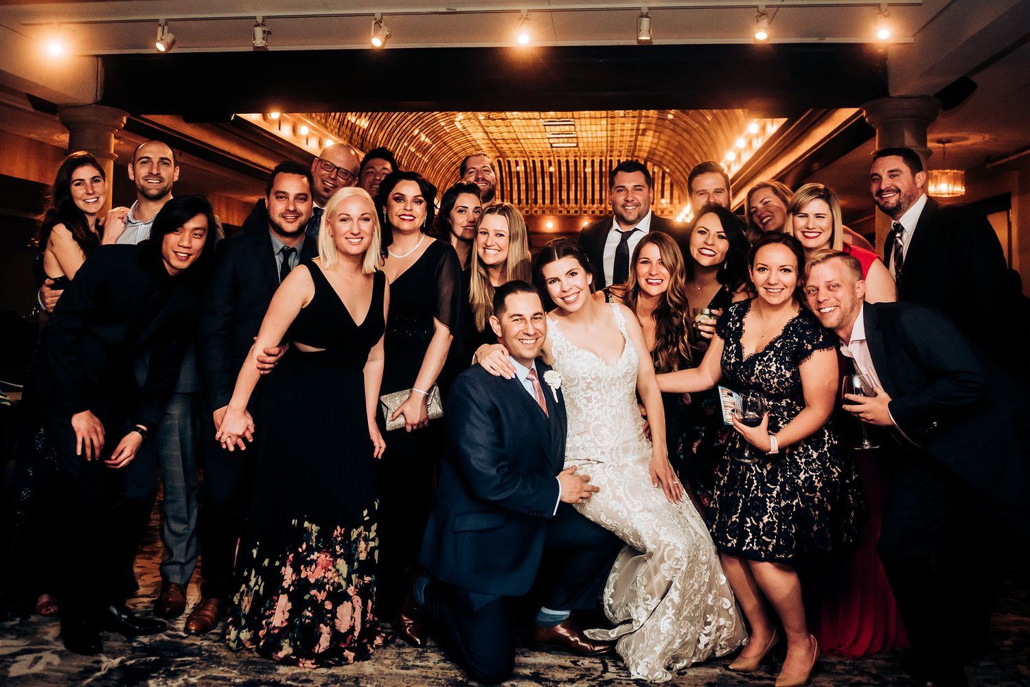 Wedding party group poses together in formal attire at an indoor reception venue with warm lighting.