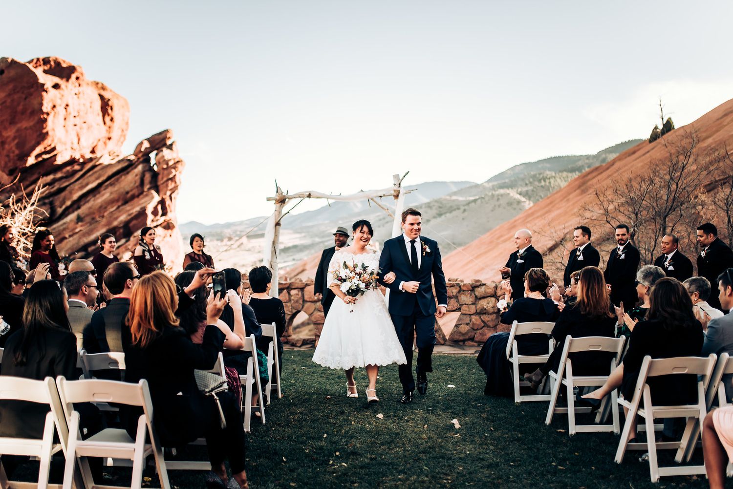 Newlyweds walk down aisle at outdoor ceremony with red rocks and white chairs against mountain backdrop.