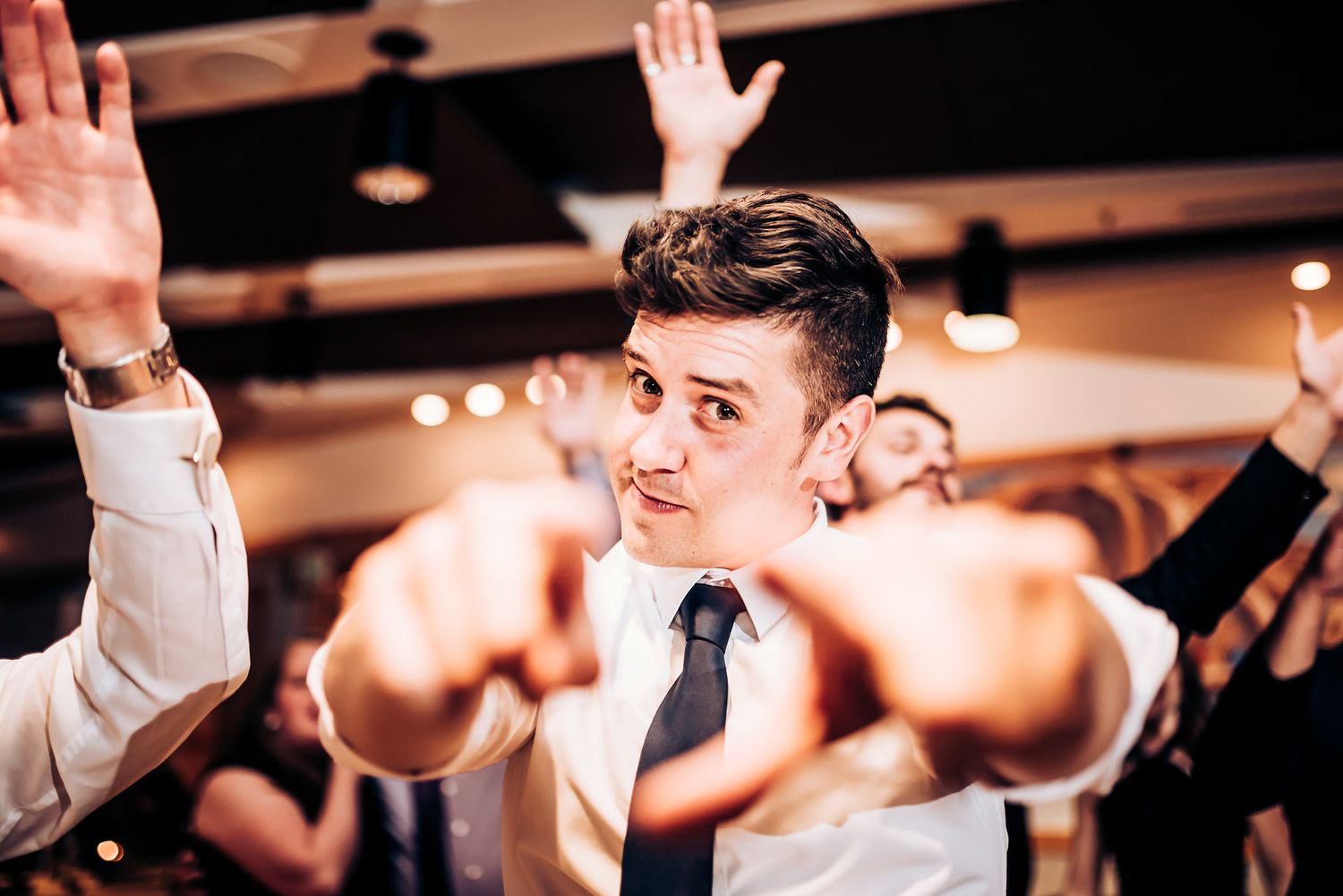 Party guest in formal attire raises hands while dancing at indoor celebration.