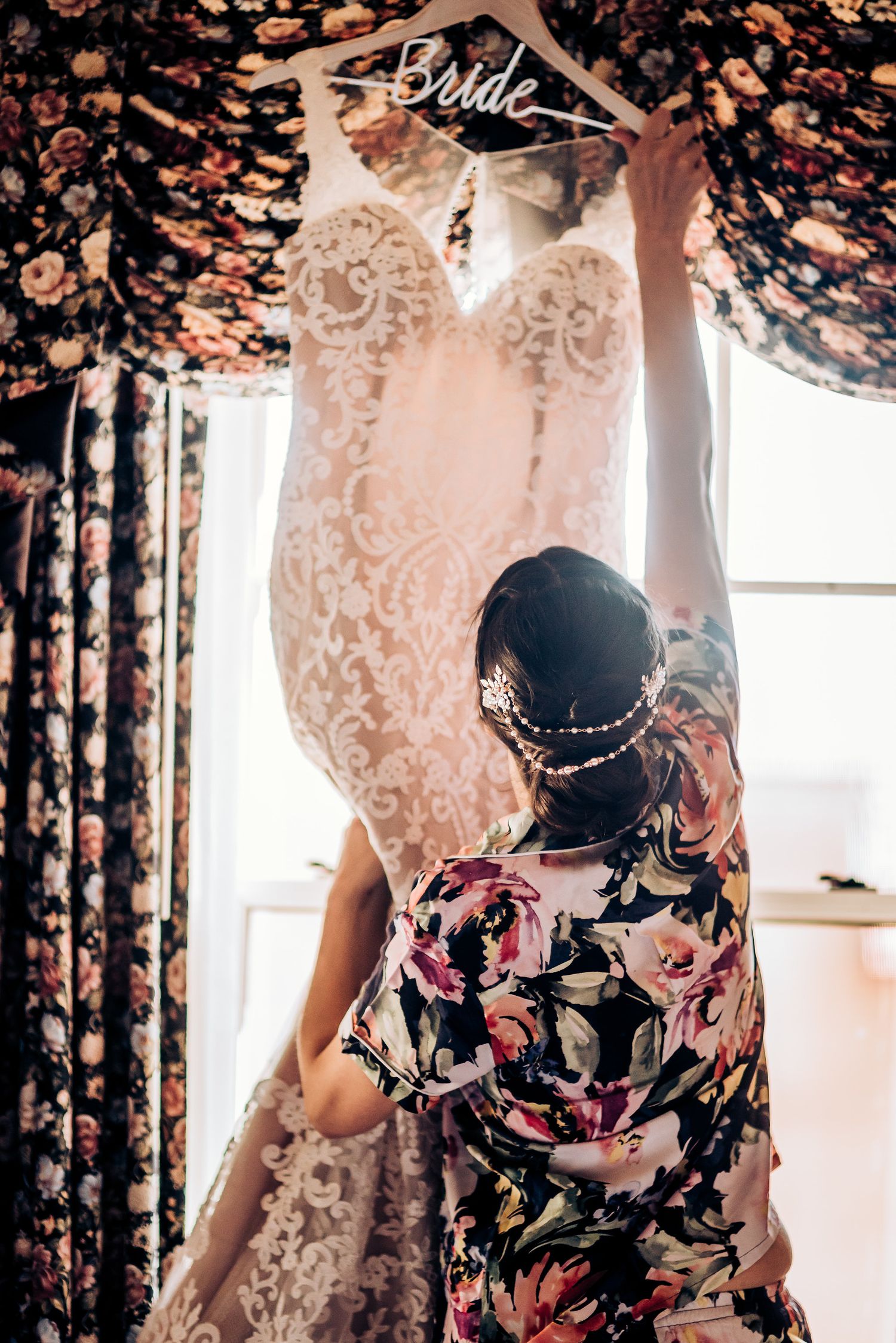 A delicate lace wedding dress hangs on display while being arranged against floral curtains.