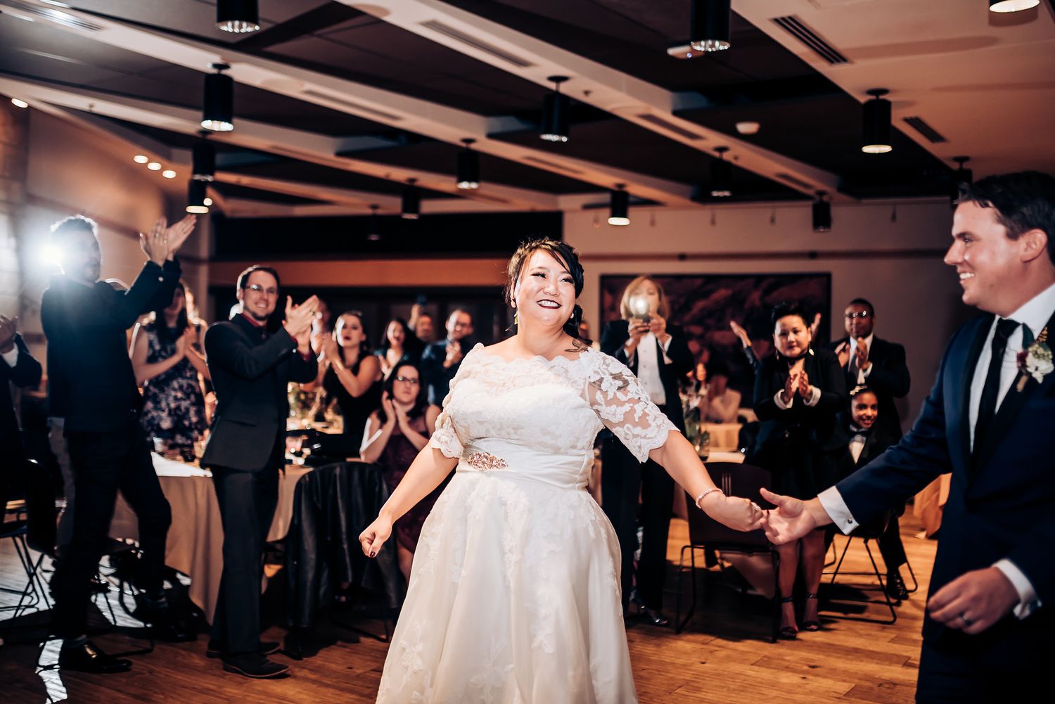 A happy couple twirls and dances in a lively reception venue surrounded by joyful wedding guests.