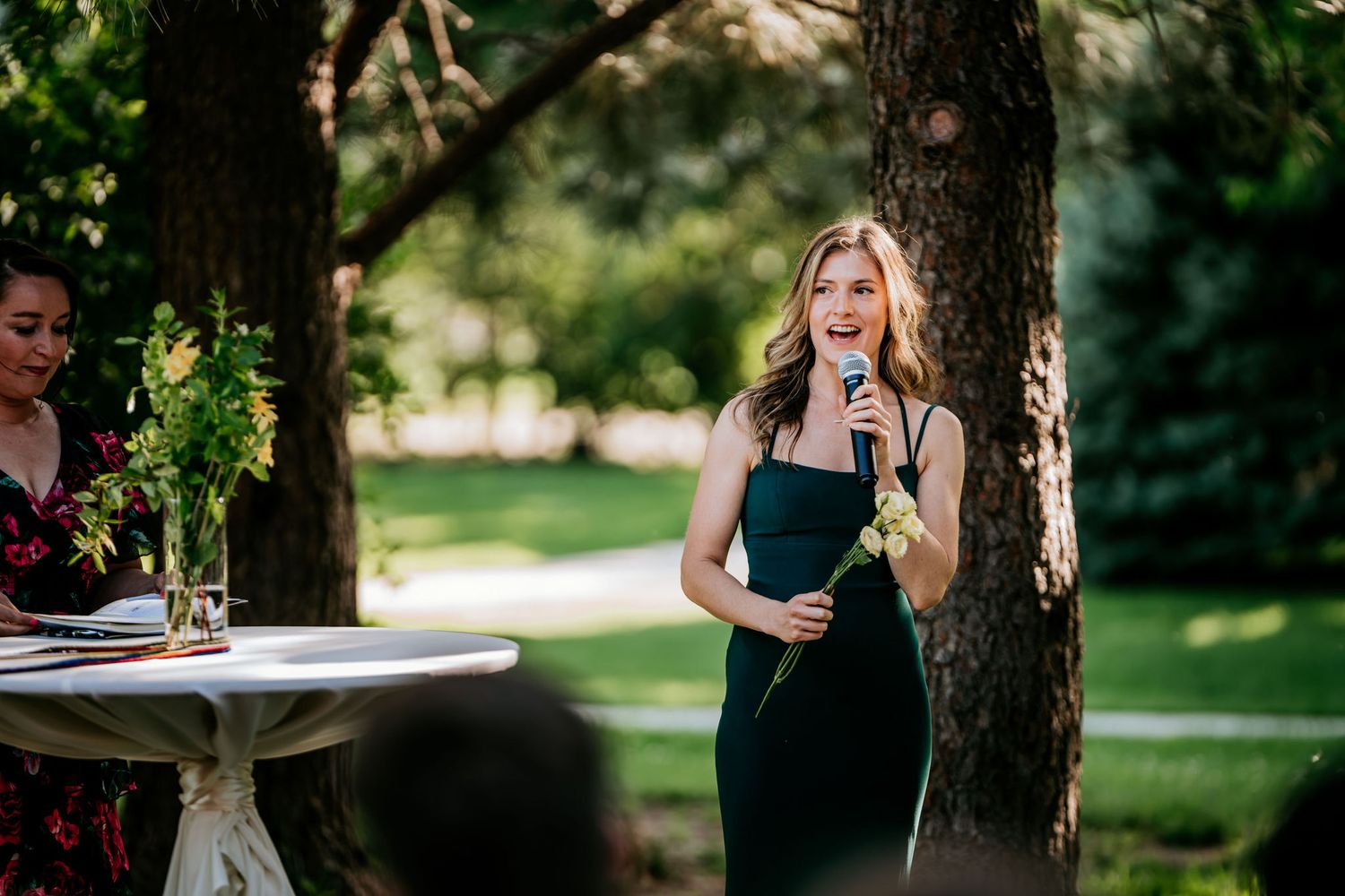 An elegant speaker in a black dress holds a microphone during an outdoor event.