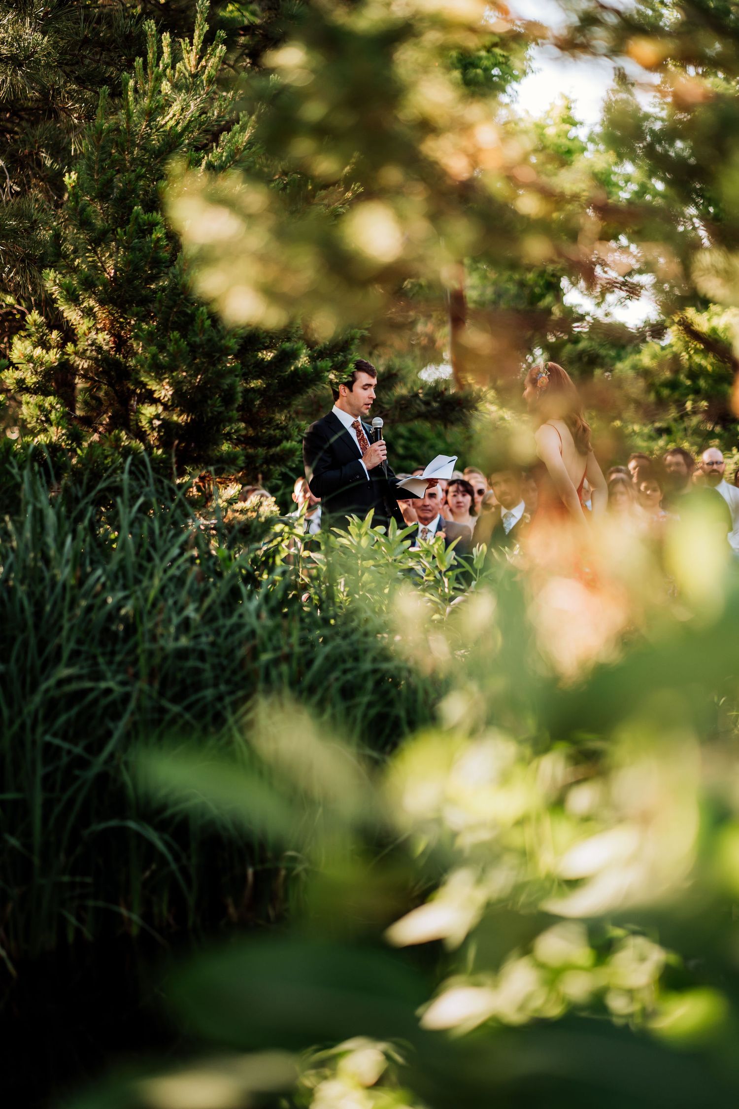 Outdoor wedding ceremony through foliage with guests seated among trees.