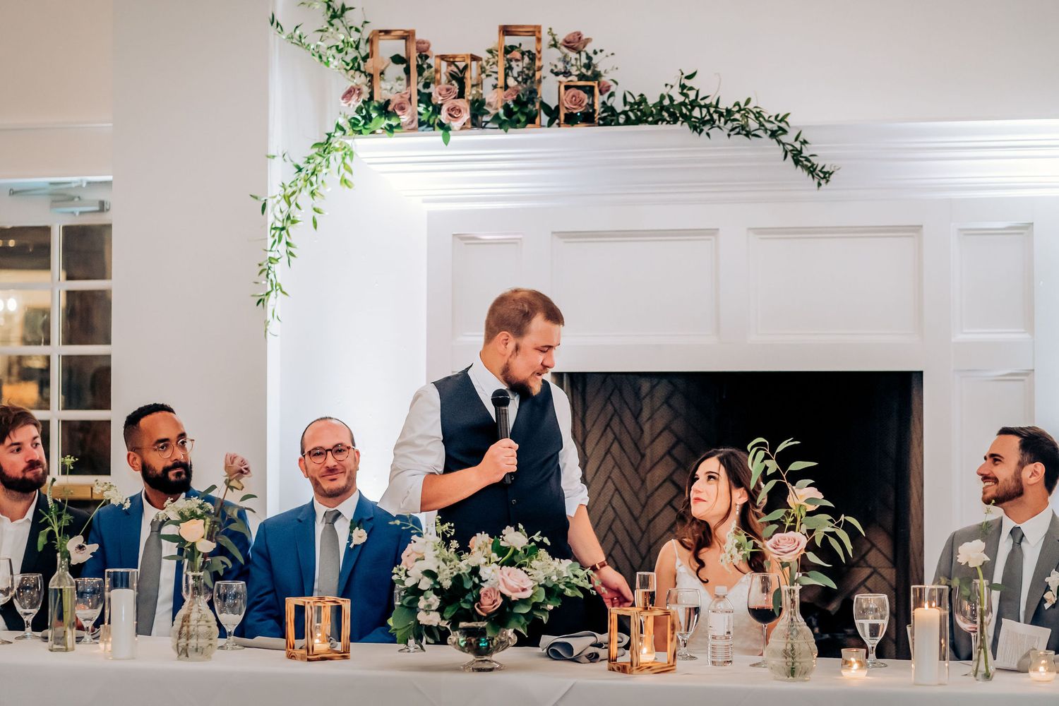 Wedding party members enjoy toasts and speeches at elegantly decorated head table with greenery.