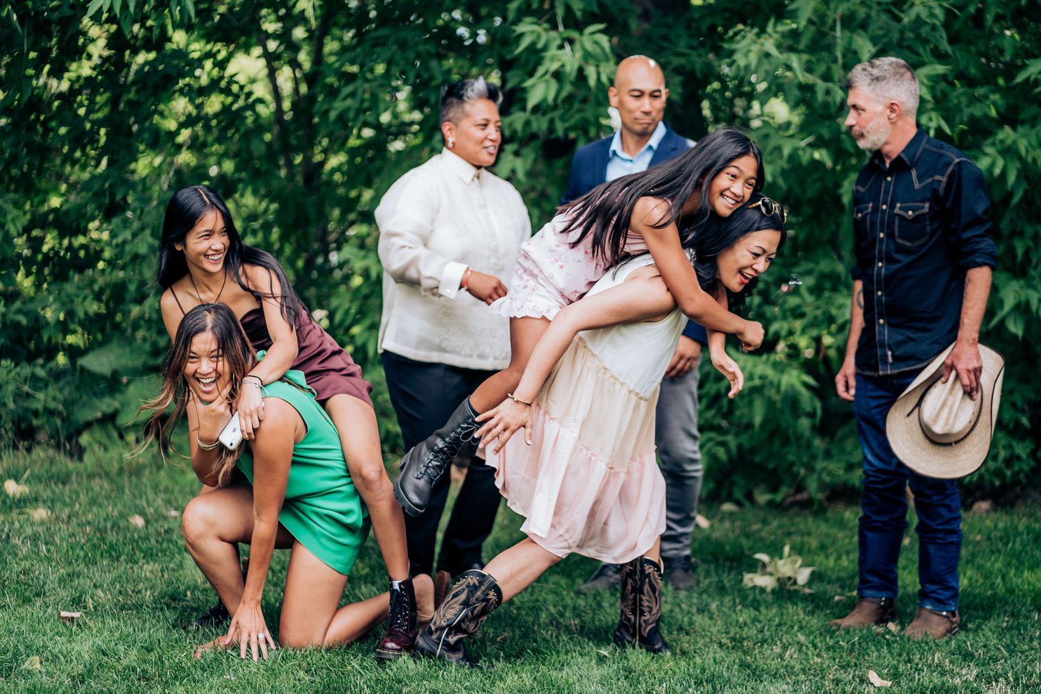 Candid moment of wedding guests playing and laughing together on a lawn with trees in background.